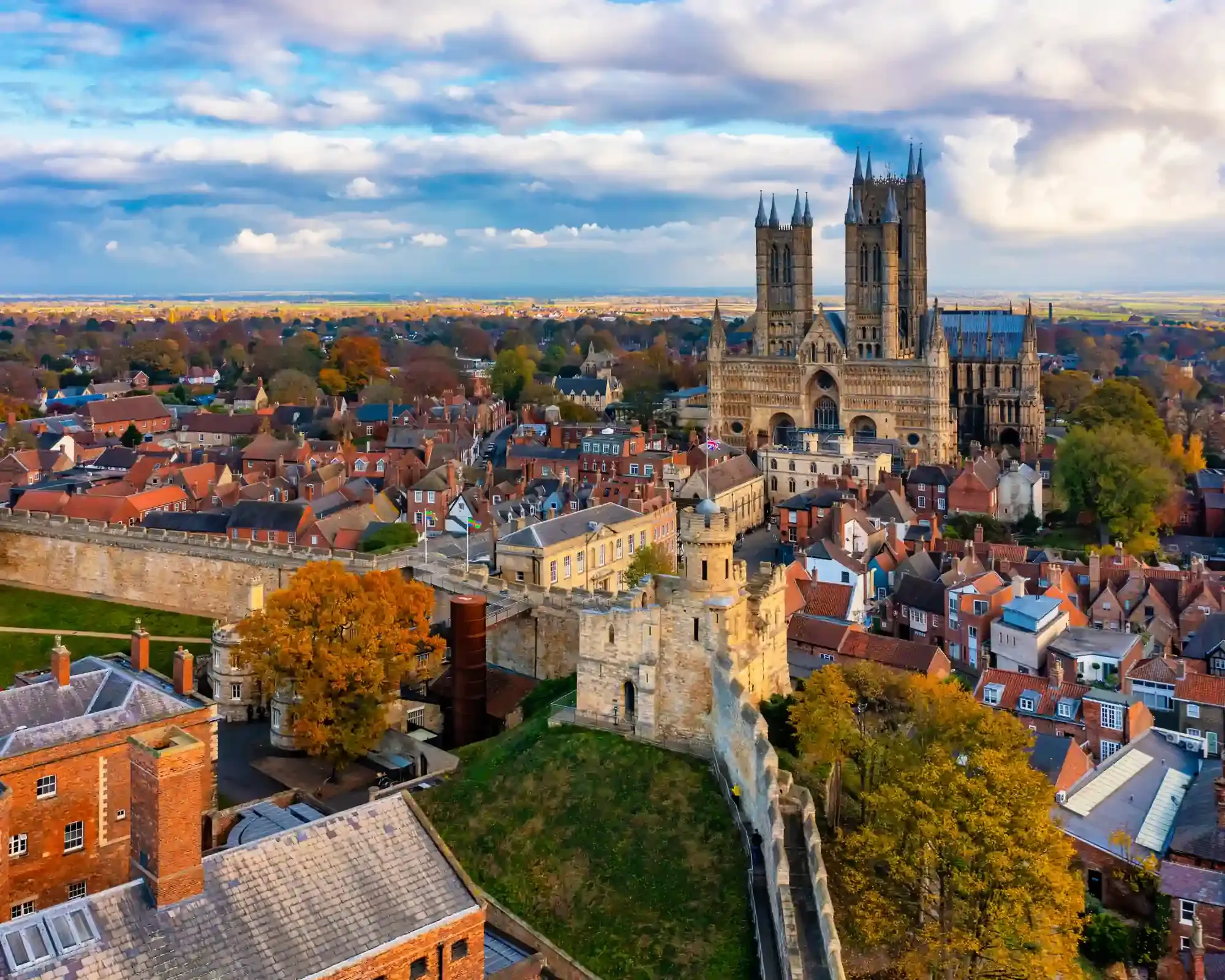 Aerial view of Lincoln with the castle walls and autumn trees in the foreground and Lincoln Cathedral rising above the red-brick rooftops.