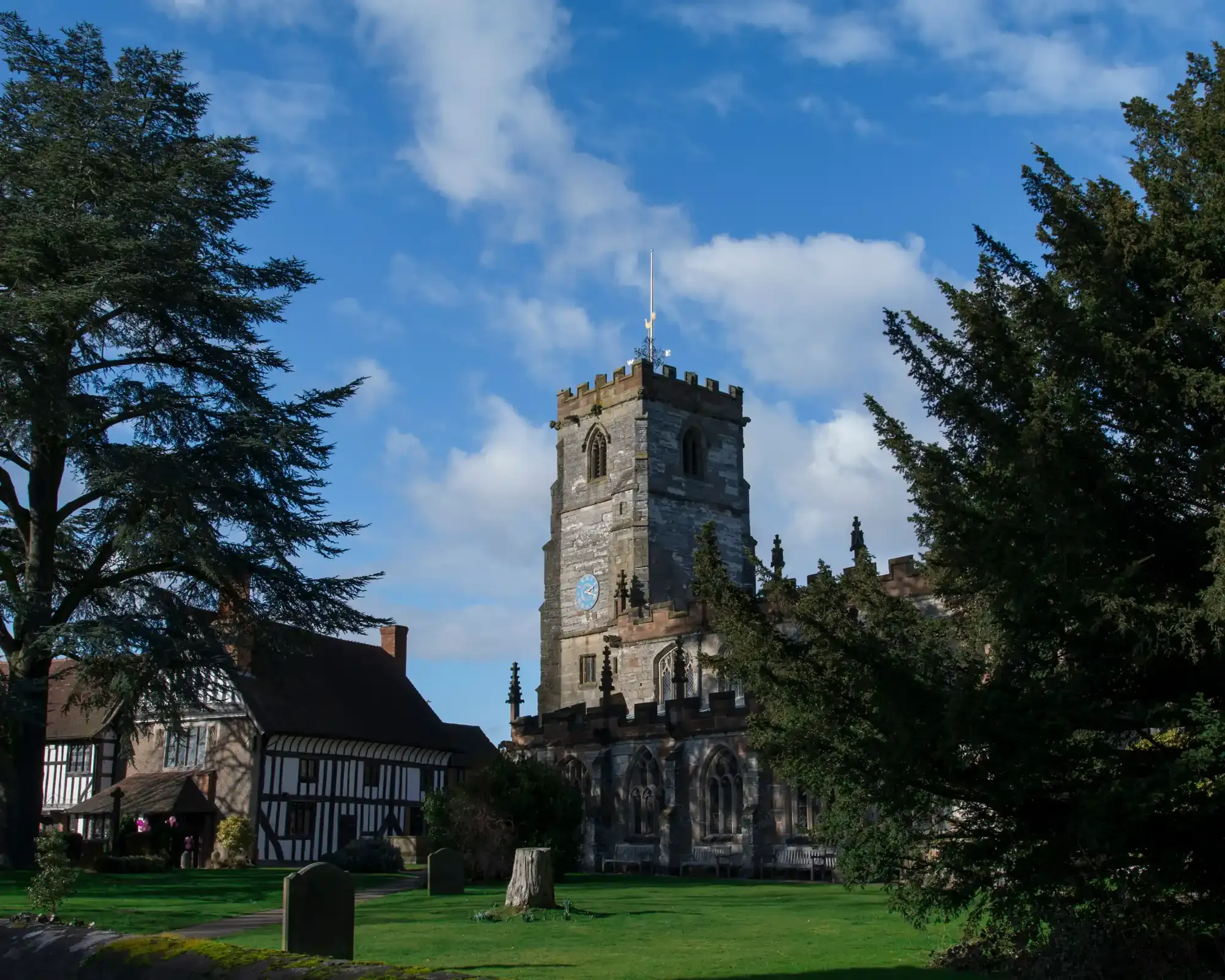 Knowle parish church tower and the timber-framed Guild House beside the churchyard, framed by tall evergreen trees in Knowle, Solihull.