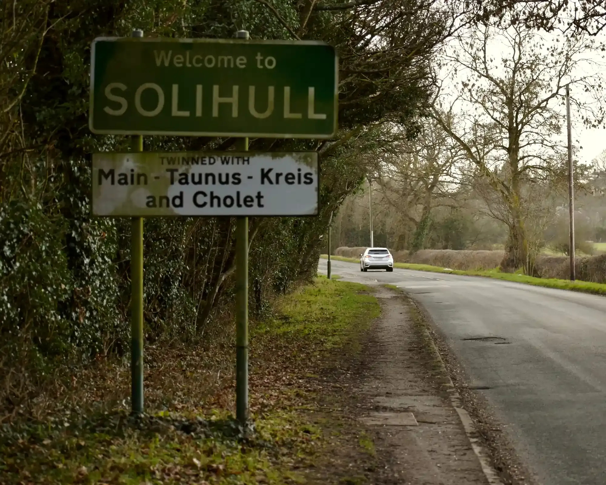 Welcome to Solihull" road sign, twinned with Main-Taunus-Kreis and Cholet, beside a quiet tree-lined country road with a car in the distance.