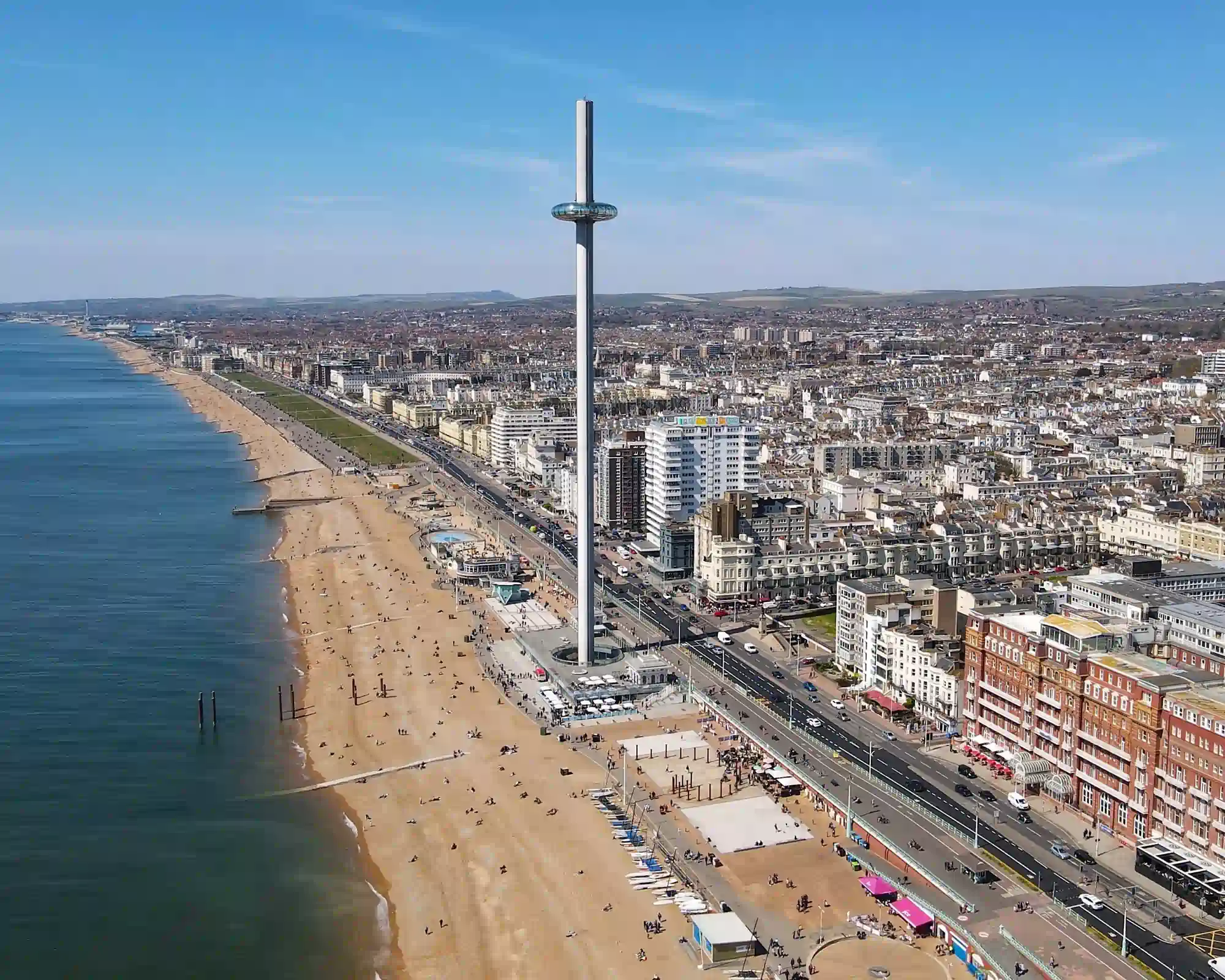 Aerial view of Brighton seafront on a sunny day, with the tall British Airways i360 viewing tower beside a sandy beach, the promenade and seafront hotels.