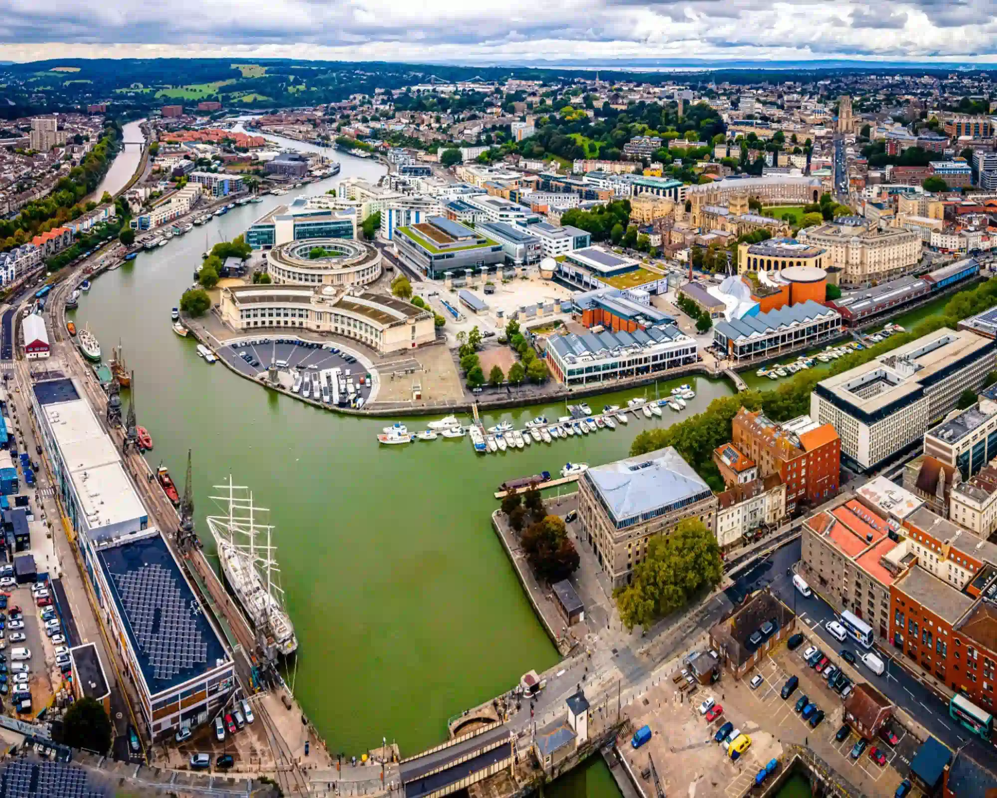 Aerial view of Bristol Harbourside, with a tall ship moored at the quay, a marina full of boats and the River Avon winding through the city.