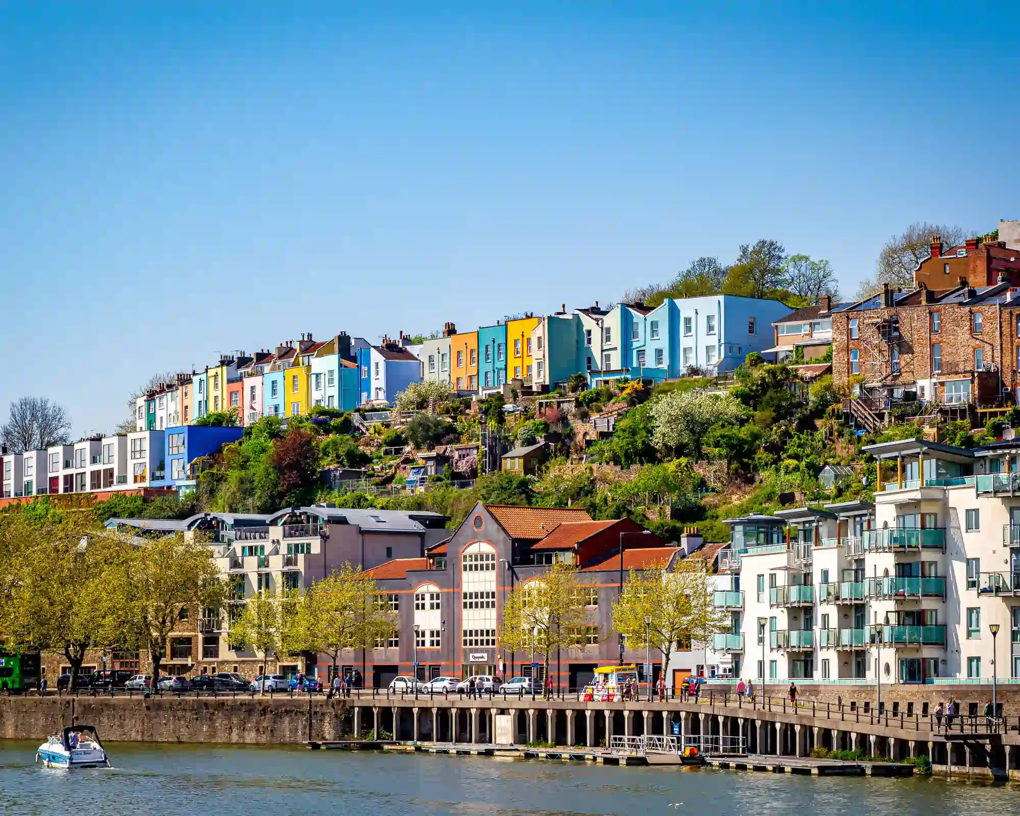 Rows of colourful painted houses on a green hillside in Cliftonwood, Bristol, above the harbourside with apartments and a boat on the water.