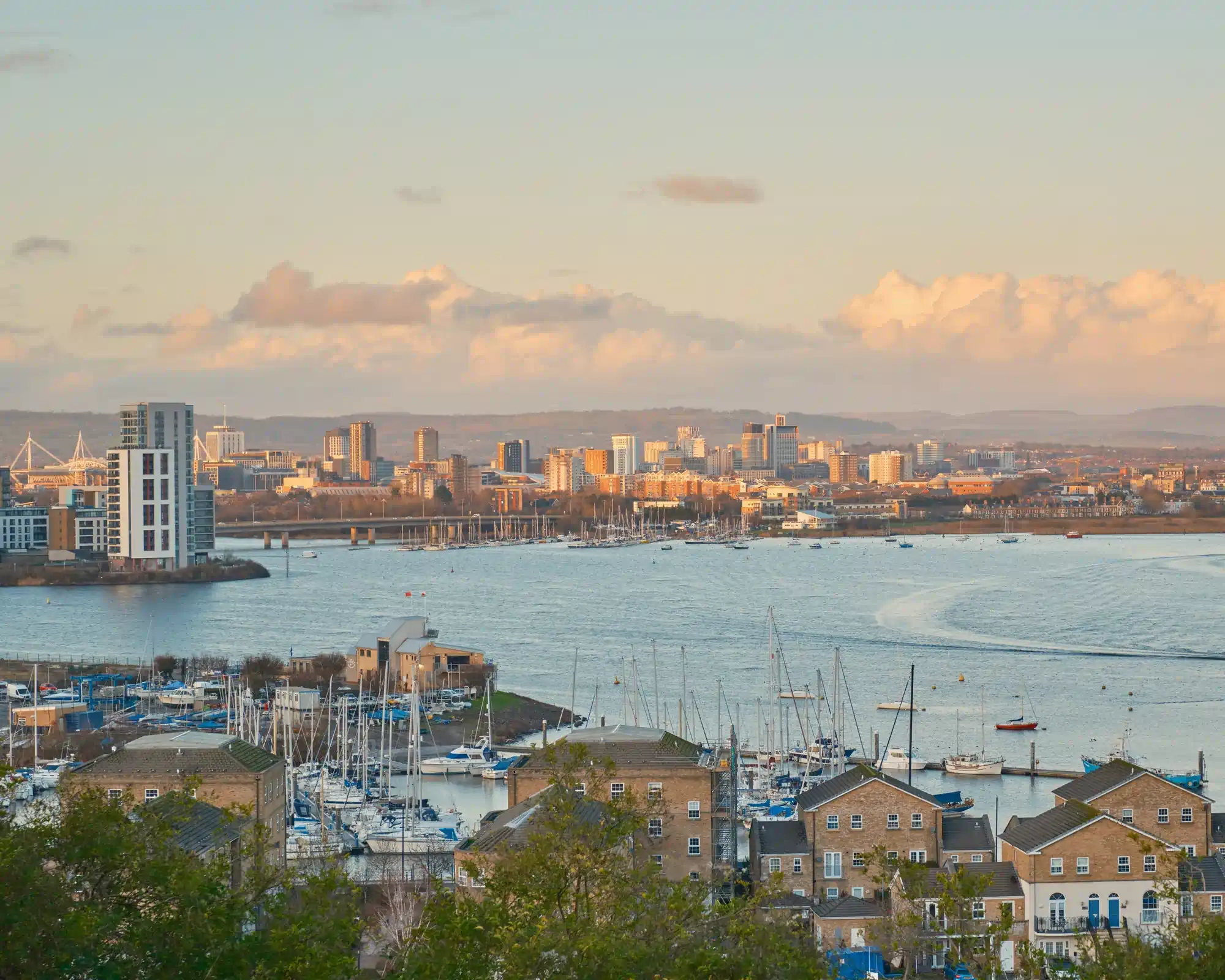Cardiff city skyline and the Principality Stadium at golden hour, seen across Cardiff Bay from Penarth, with a marina full of sailing boats and brick townhouses in the foreground.