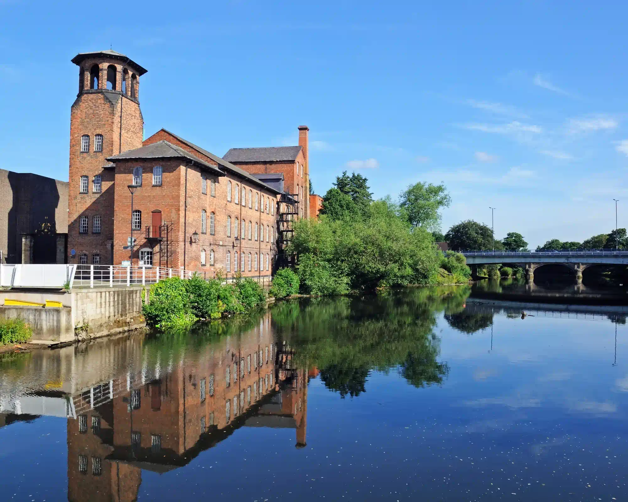 Derby Silk Mill, a red-brick mill with a tall bell tower, reflected in the still River Derwent on a sunny day, with trees and a road bridge beyond.