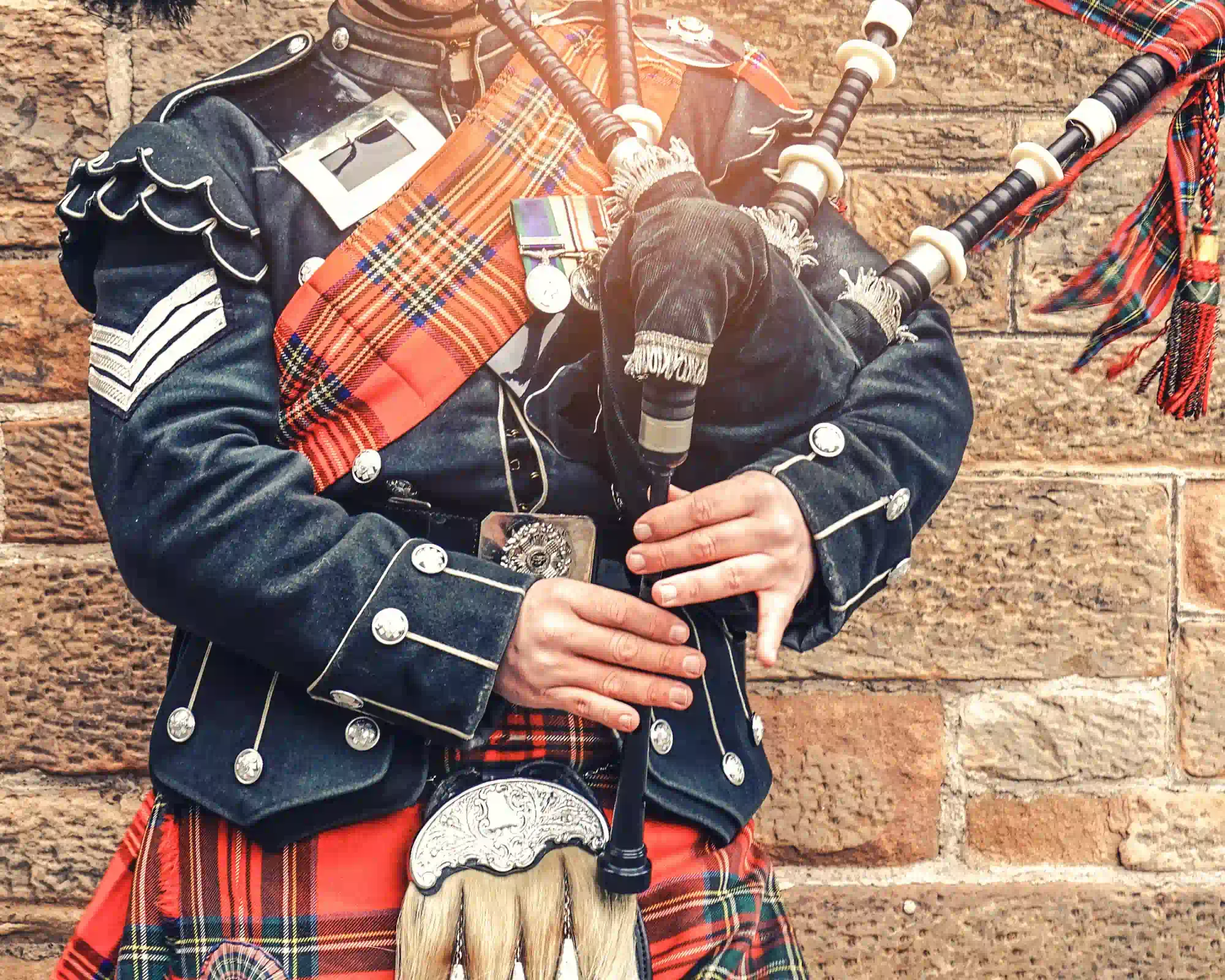 A piper in a Highland military uniform and red tartan kilt playing the bagpipes against a sandstone wall.