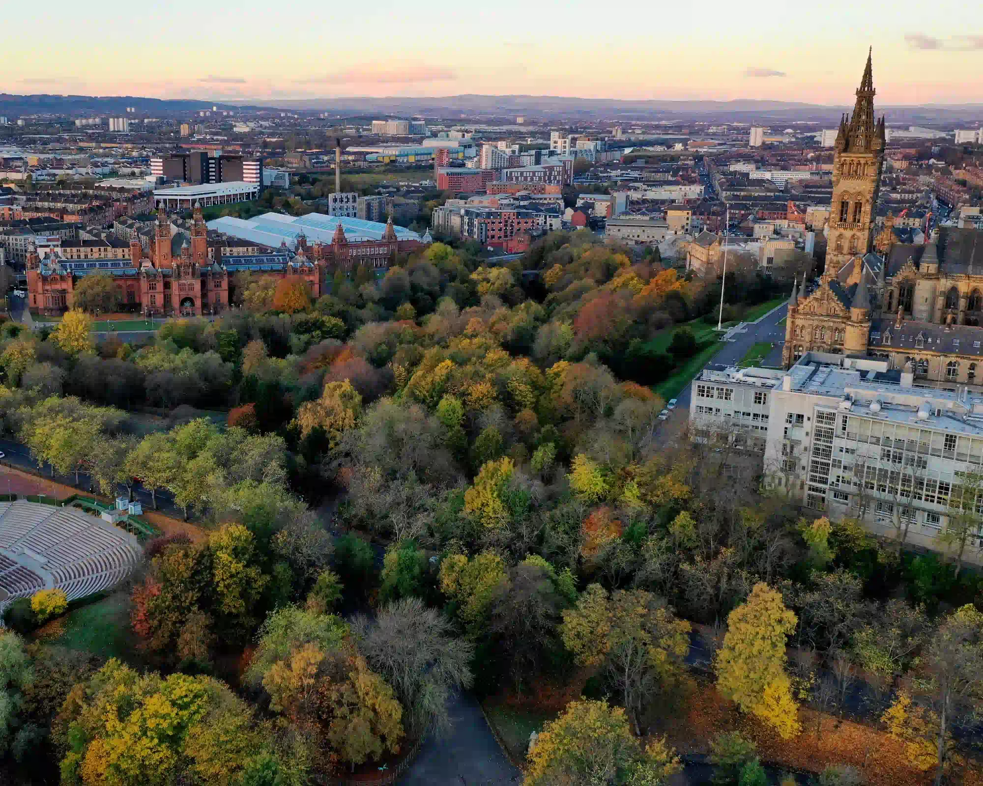 Aerial view of Kelvingrove Park in Glasgow in autumn, with Kelvingrove Art Gallery and Museum and the University of Glasgow's Gothic tower among colourful trees at sunset.