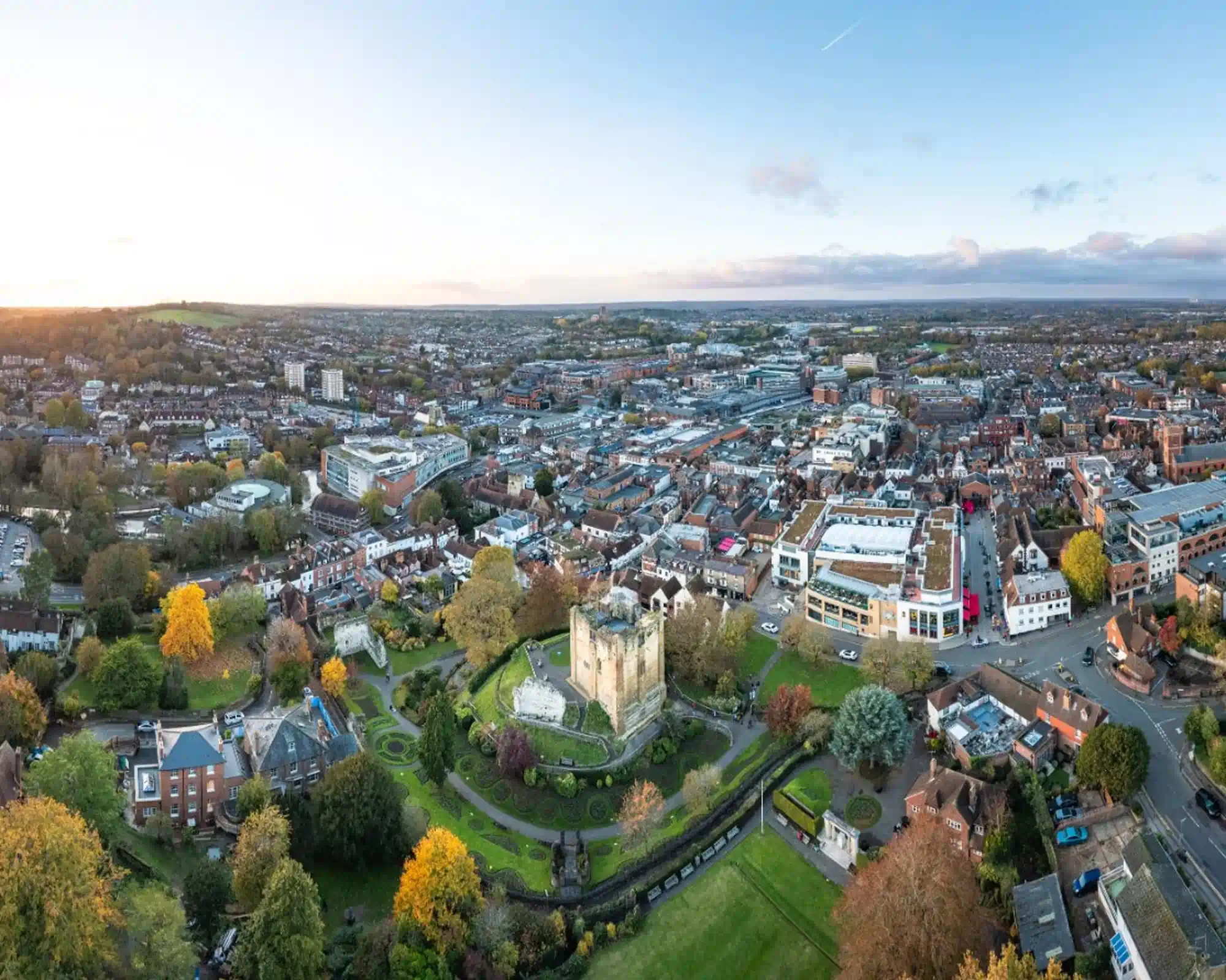 Aerial view of Guildford at sunset in autumn, with the Norman castle keep in its gardens surrounded by golden trees, and the town centre's rooftops stretching beyond.