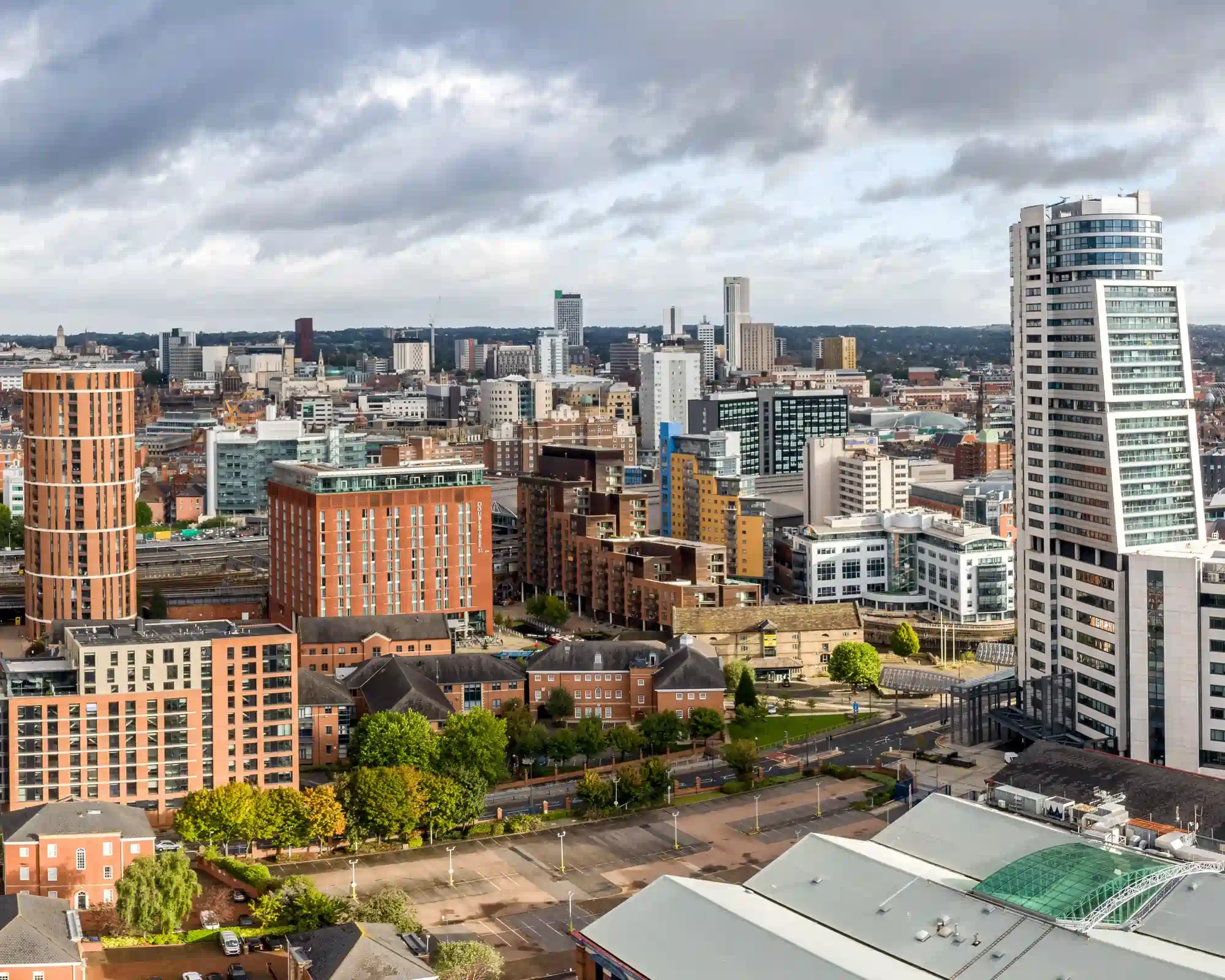 Leeds city centre skyline under a dramatic cloudy sky, with the tall Bridgewater Place tower, a round red-brick apartment tower and office blocks stretching into the distance.