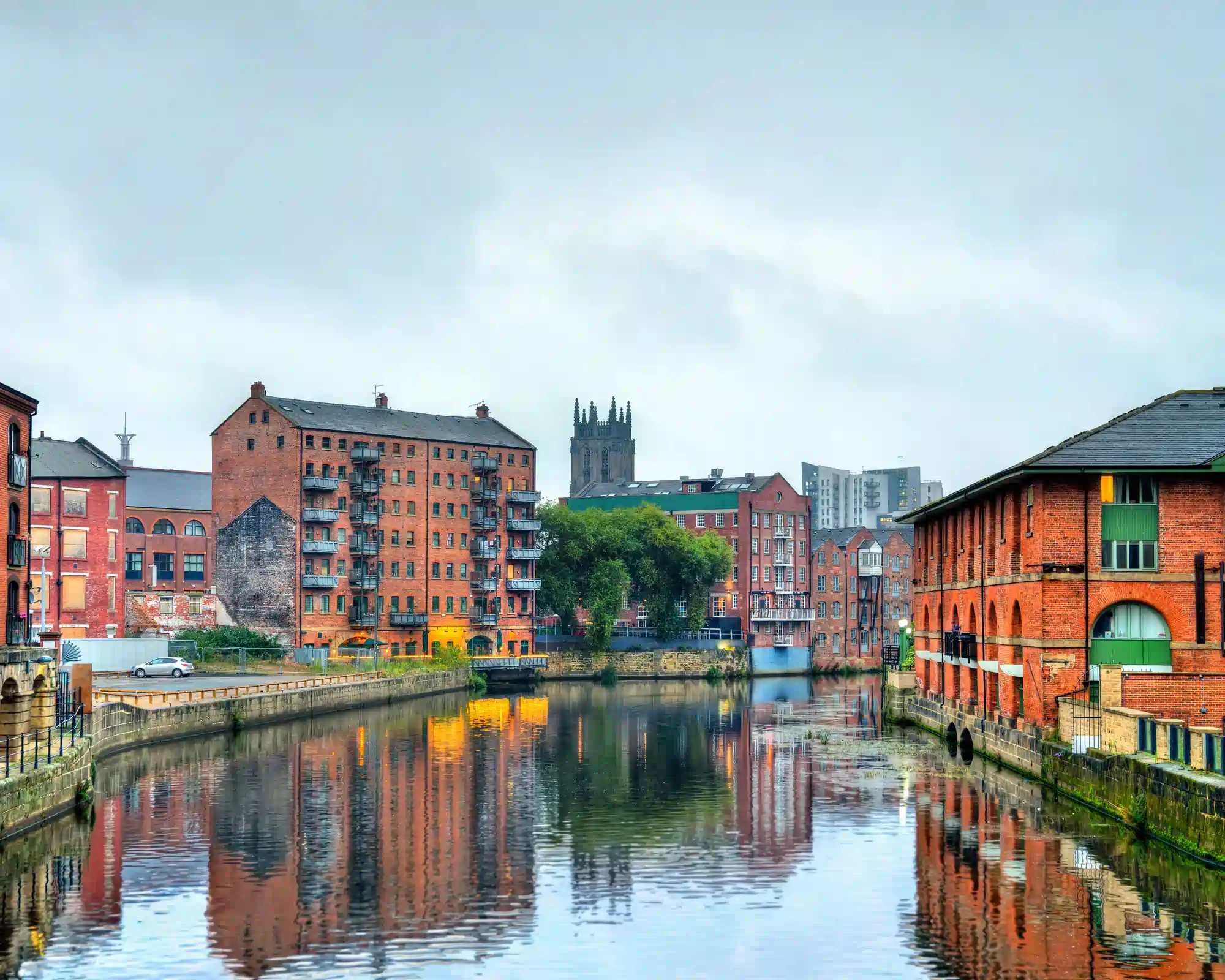 Red-brick former warehouses along the River Aire in Leeds, reflected in the still water, with the tower of Leeds Minster rising behind under a grey sky.