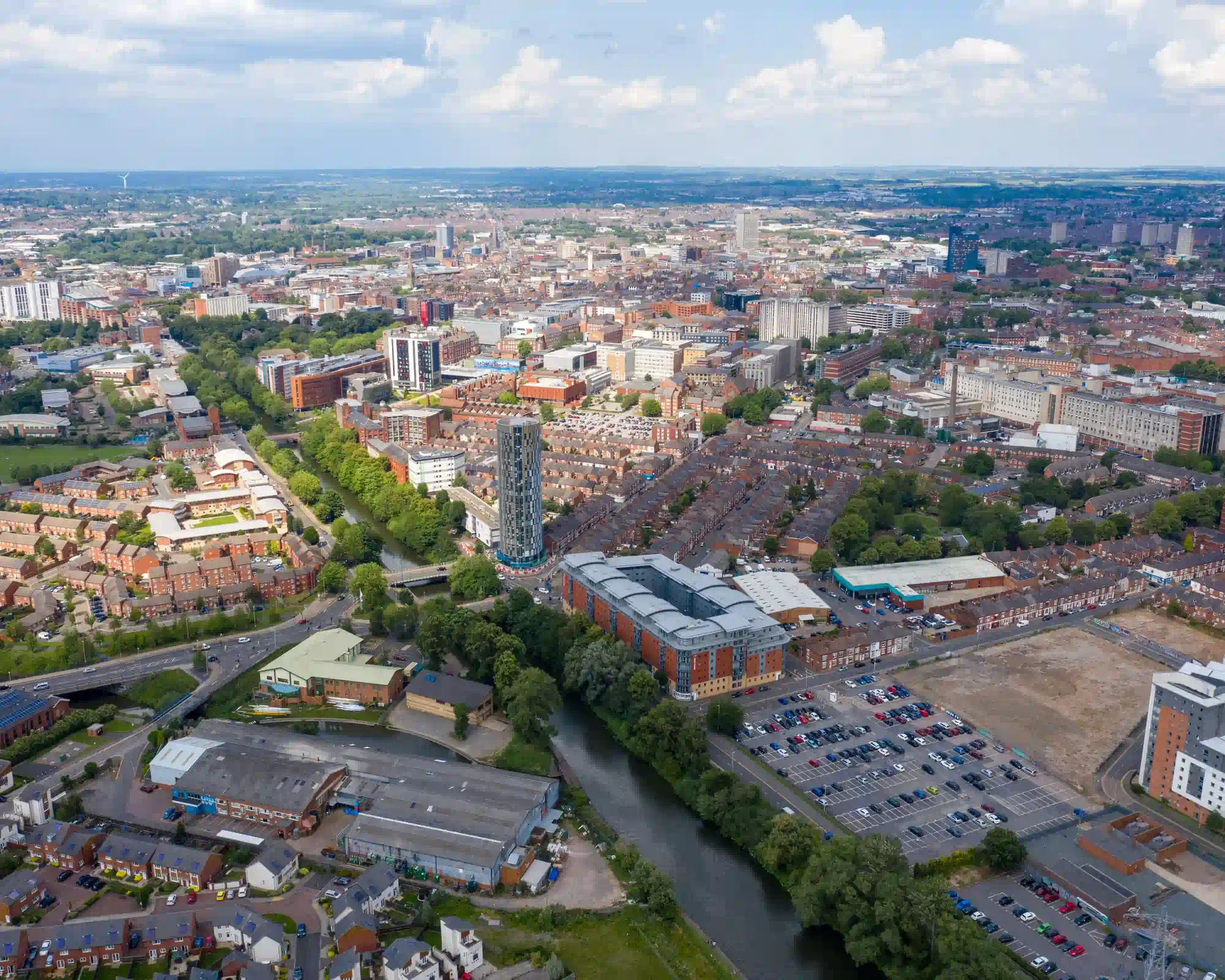 Aerial view of Leicester on a sunny day, with the River Soar winding past a tall round tower, rows of red-brick terraced houses and the city centre beyond.