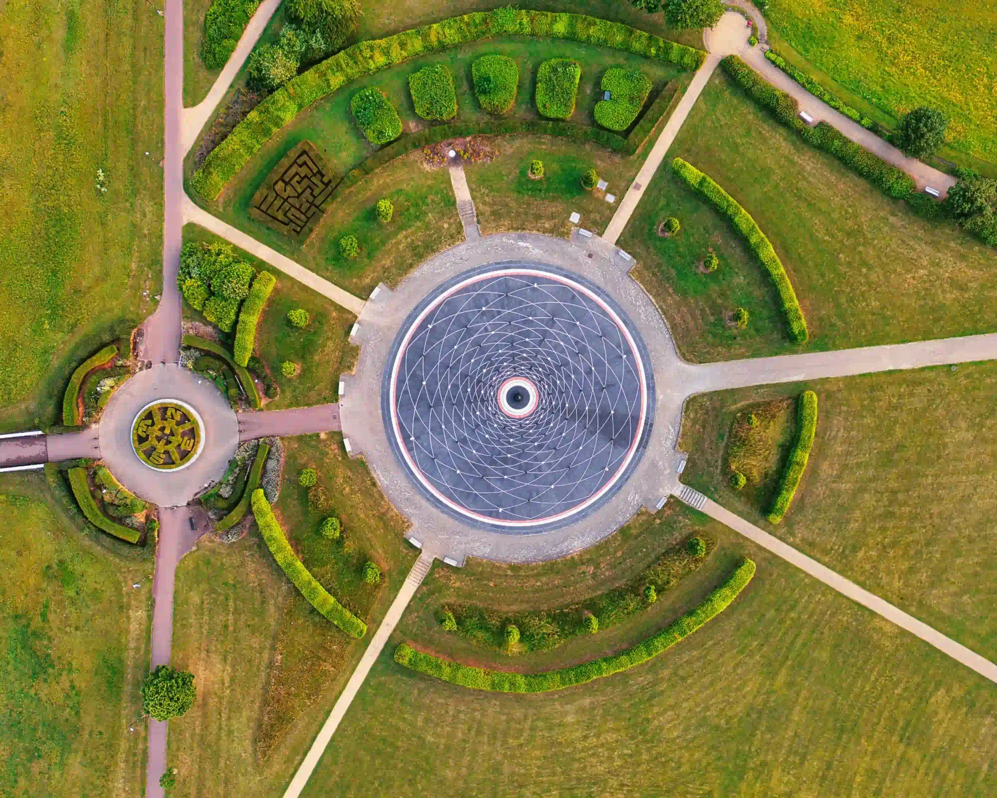 Overhead view of a circular paved plaza with a geometric pattern in a Milton Keynes park, surrounded by curved hedges, a small maze, a compass garden and paths across the grass.