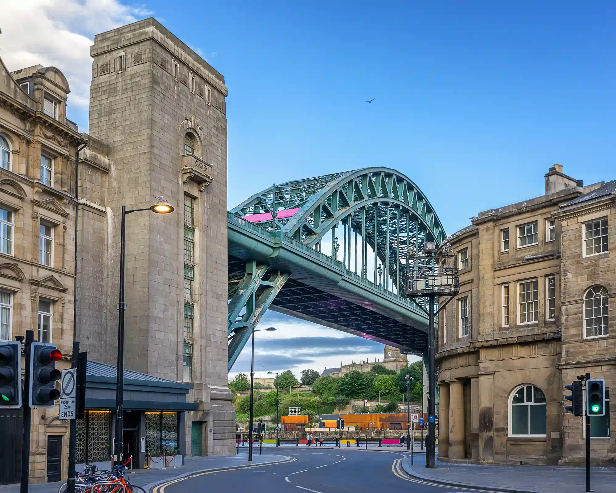 The green steel arch of the Tyne Bridge in Newcastle, framed by historic sandstone buildings on the Quayside under a blue sky.