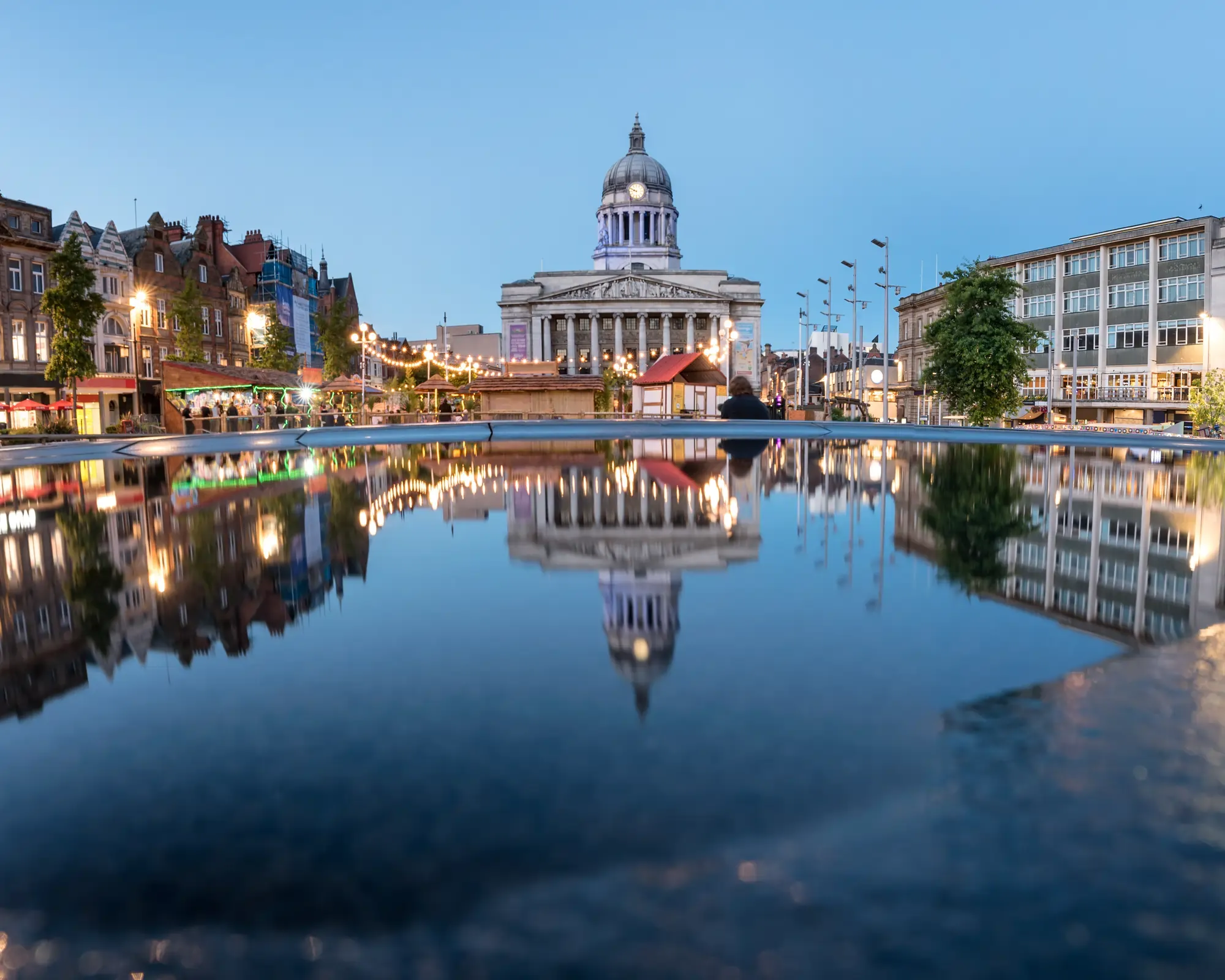 Nottingham Council House at dusk, reflected in a pool in Old Market Square, with lit-up buildings and market stalls on either side.