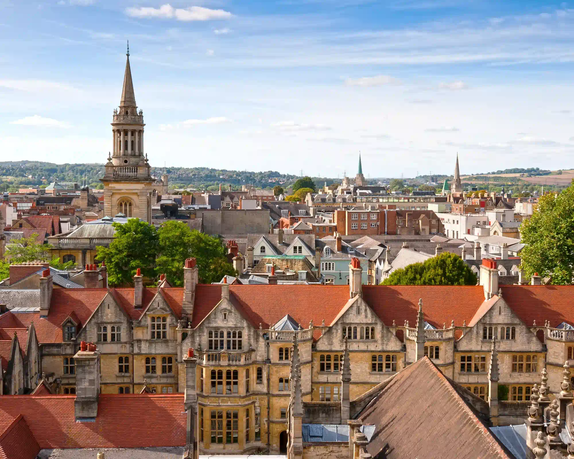 Oxford's rooftops on a sunny day, with the spire of Lincoln College Library rising above red-tiled college roofs, stone gables and distant green hills.