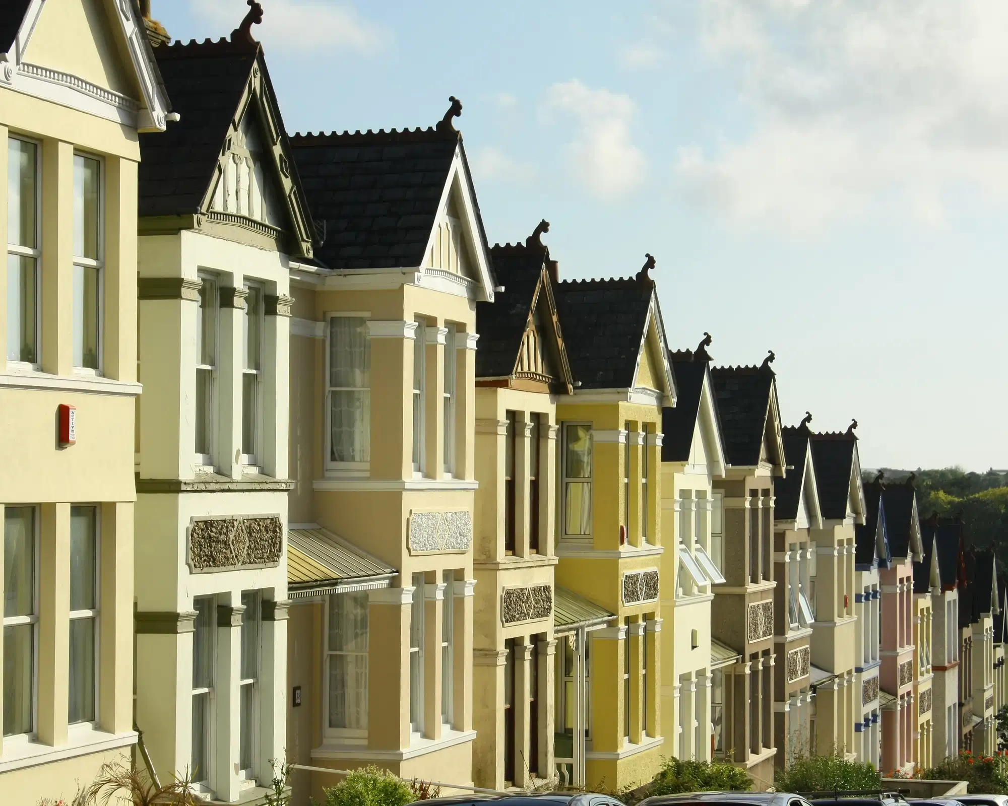 A row of Victorian terraced houses in Plymouth painted in cream and yellow, with bay windows and pointed gables, running downhill towards green trees.