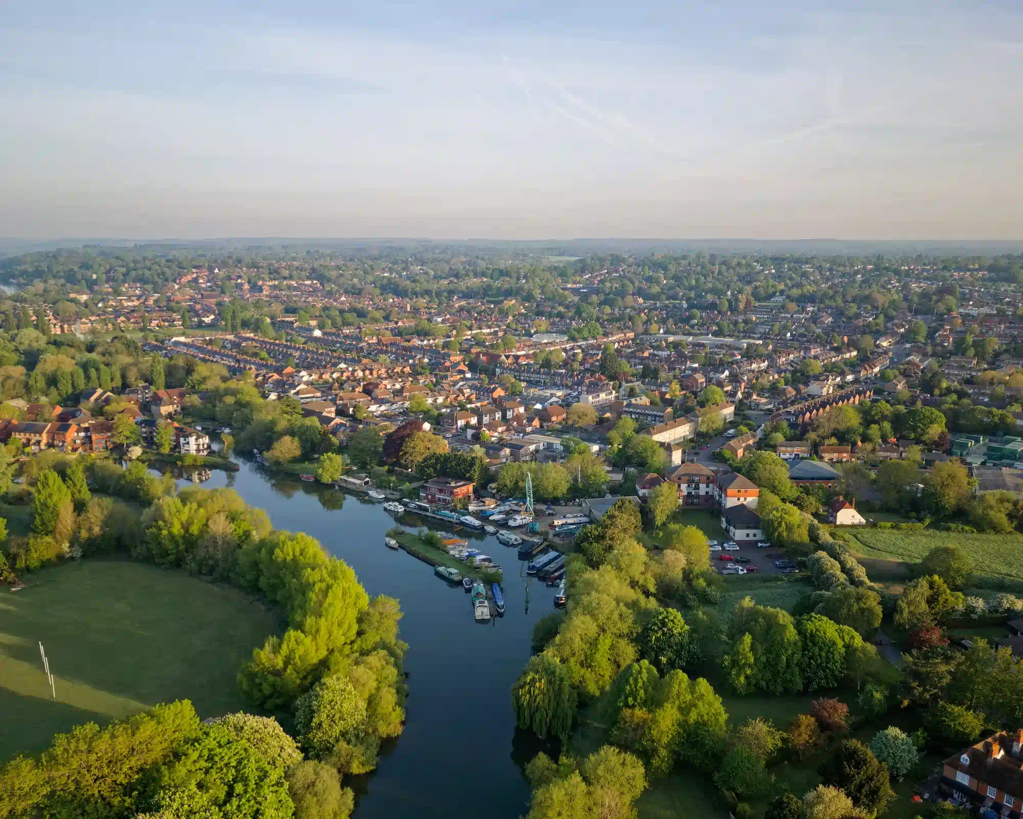 Aerial view of Reading in soft morning light, with a tree-lined river and moored boats winding past playing fields towards rows of red-brick houses.