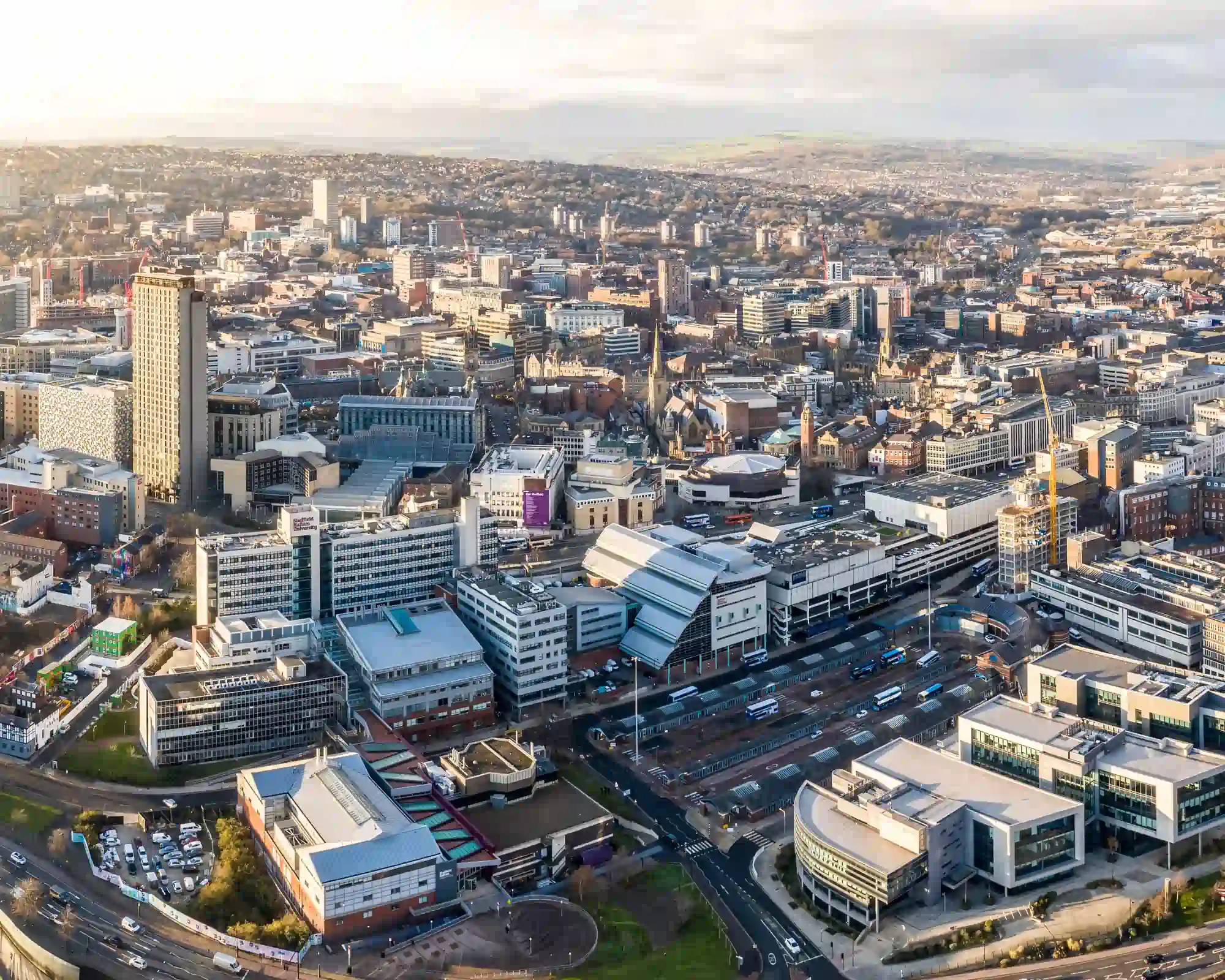 Aerial view of Sheffield city centre in golden afternoon light, with a tall tower block, church spires and office buildings, and green hills in the distance.