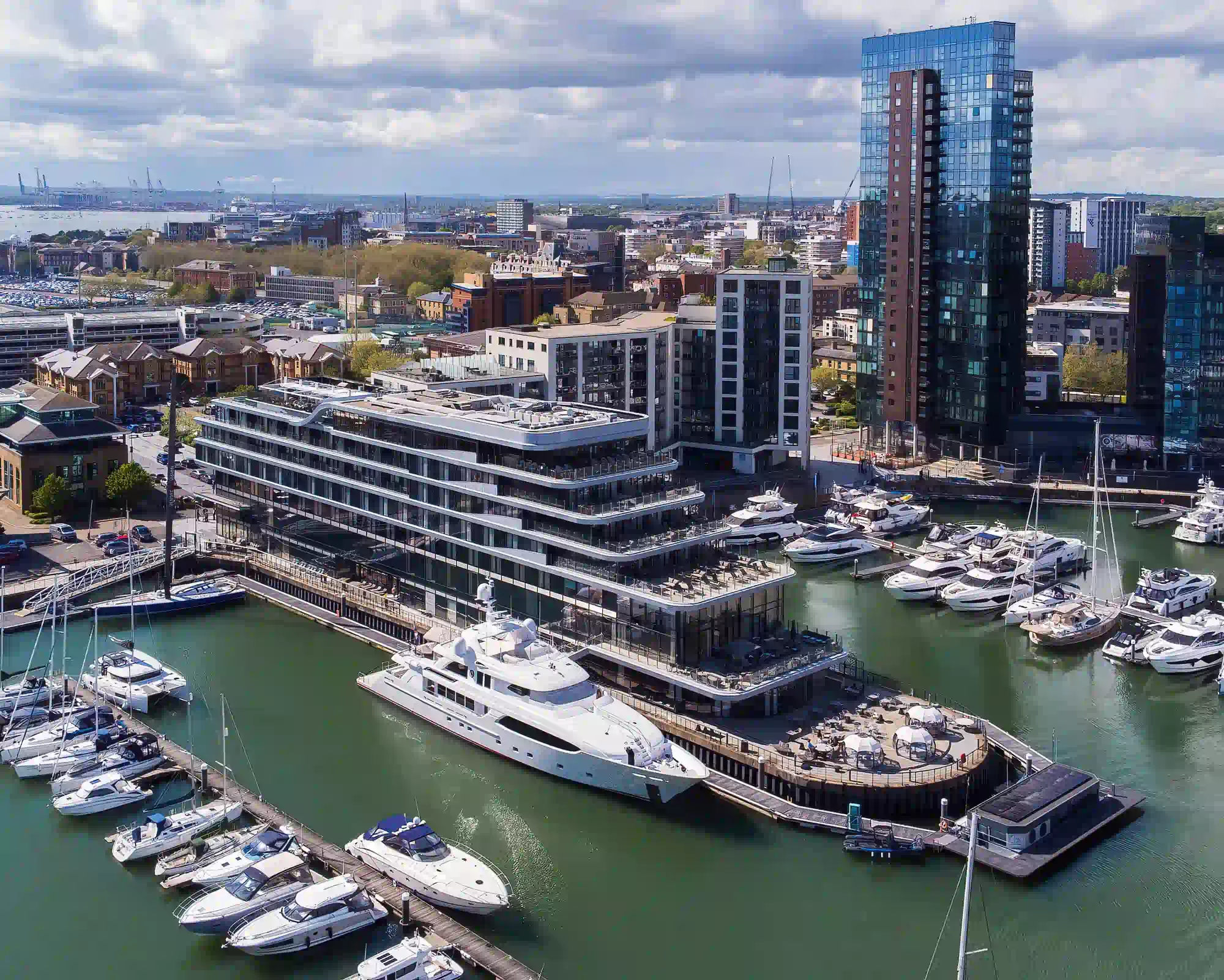 Ocean Village marina in Southampton, with a superyacht moored beside a modern waterfront hotel, rows of boats and a glass tower under a cloudy sky.