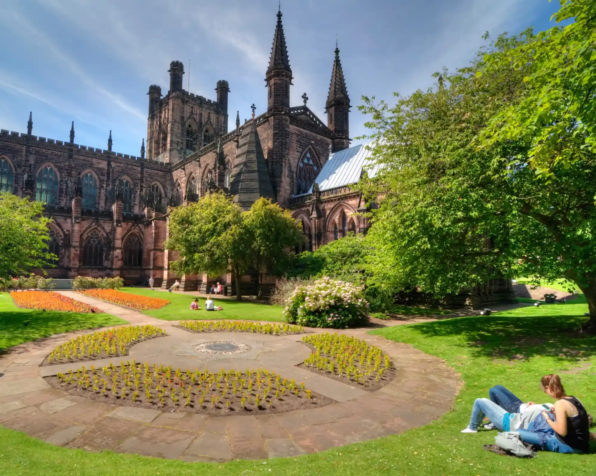 Chester Cathedral’s red sandstone tower and spires above the cathedral gardens, with flower beds and people relaxing on the lawn.