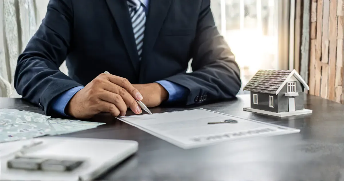 Man in suit sitting at a desk with pen and paper plus a key and a model house.