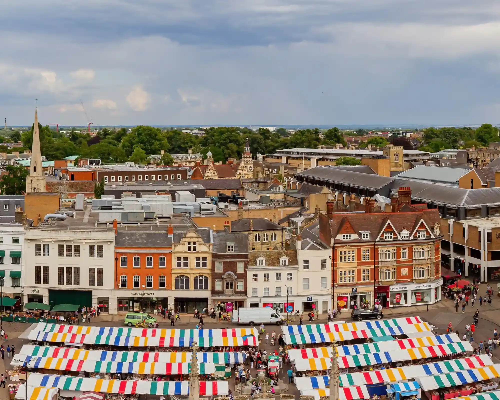 Aerial view of Cambridge Market Square with colourful striped stall awnings and historic shopfronts, seen from Great St Mary's Church.