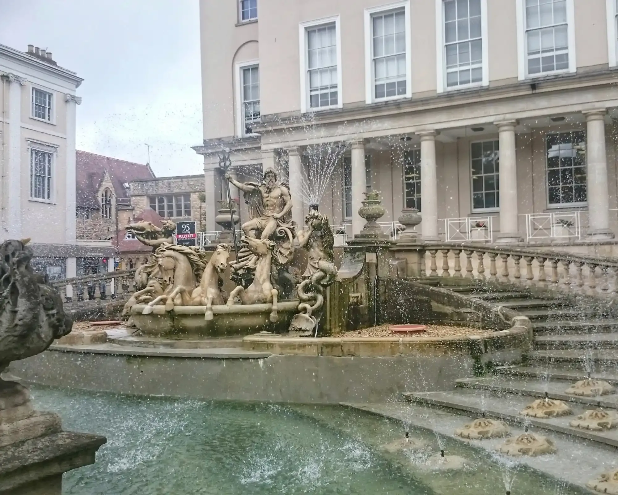 Neptune's Fountain on the Promenade in Cheltenham, with stone sea horses and water jets in front of a pale Regency building with columns.