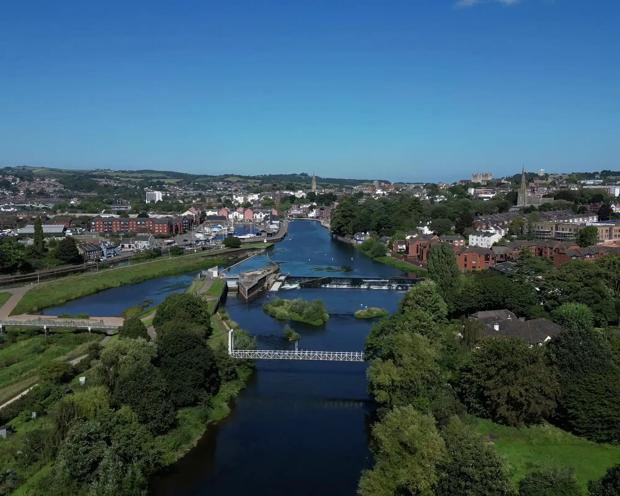 Aerial view of the River Exe in Exeter, with the Trews Weir, a suspension footbridge and the historic Quayside, and the cathedral on the skyline.