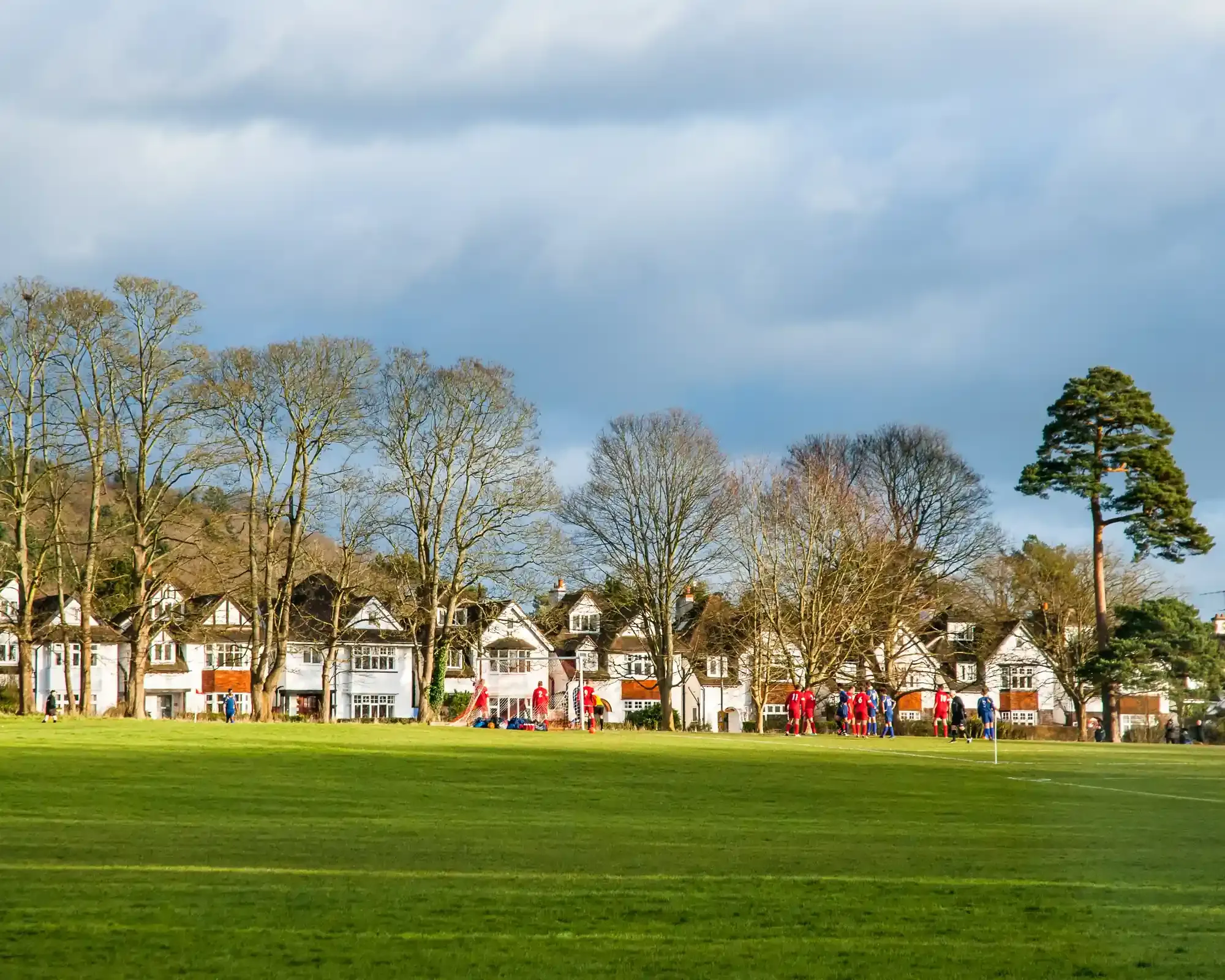 Football match on a green playing field in Guildford, in front of a row of white Arts and Crafts houses and tall trees.