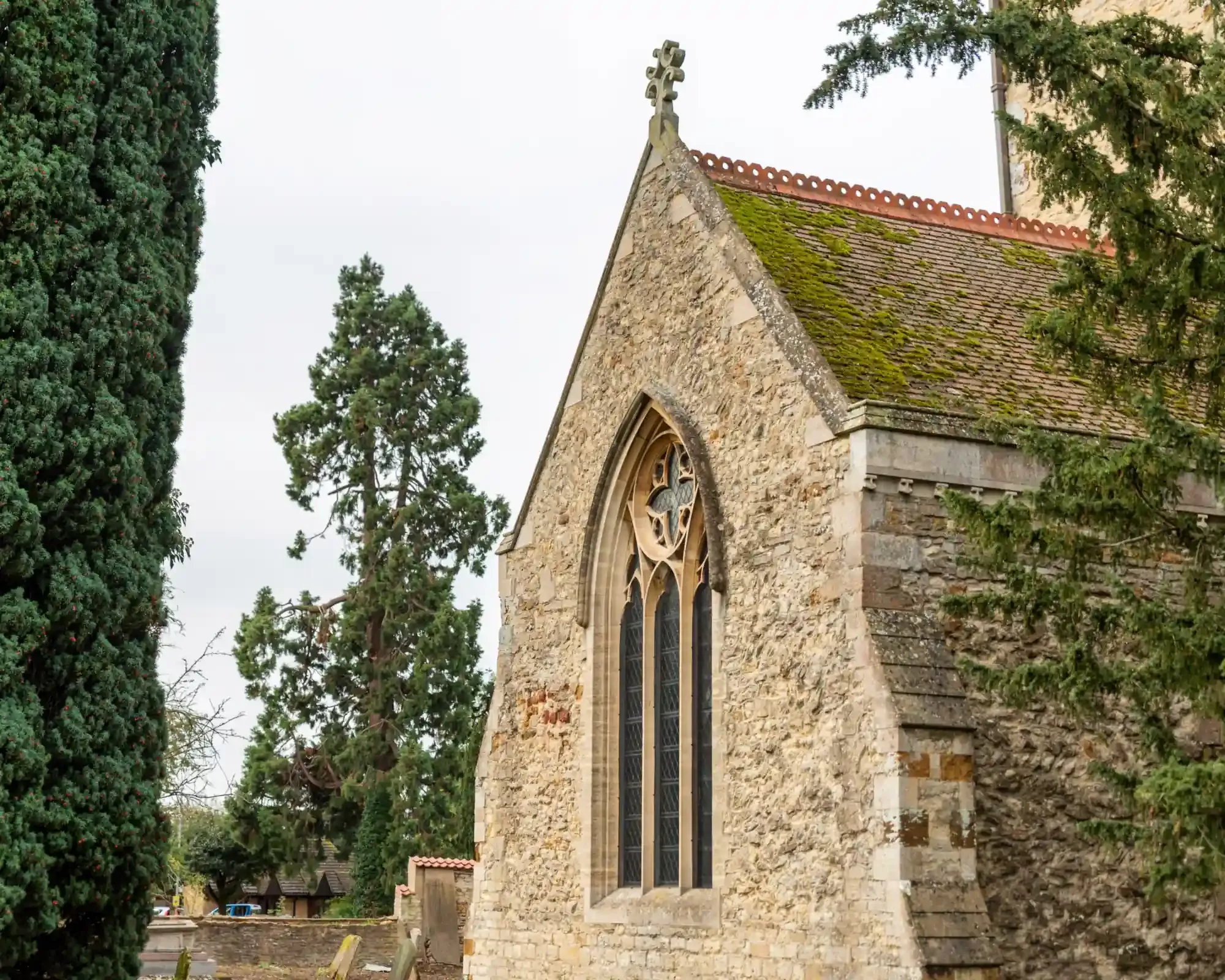 St Botolph's Church in Barton Seagrave, Kettering, with its stone gable and Gothic window framed by yew trees and a tall redwood.