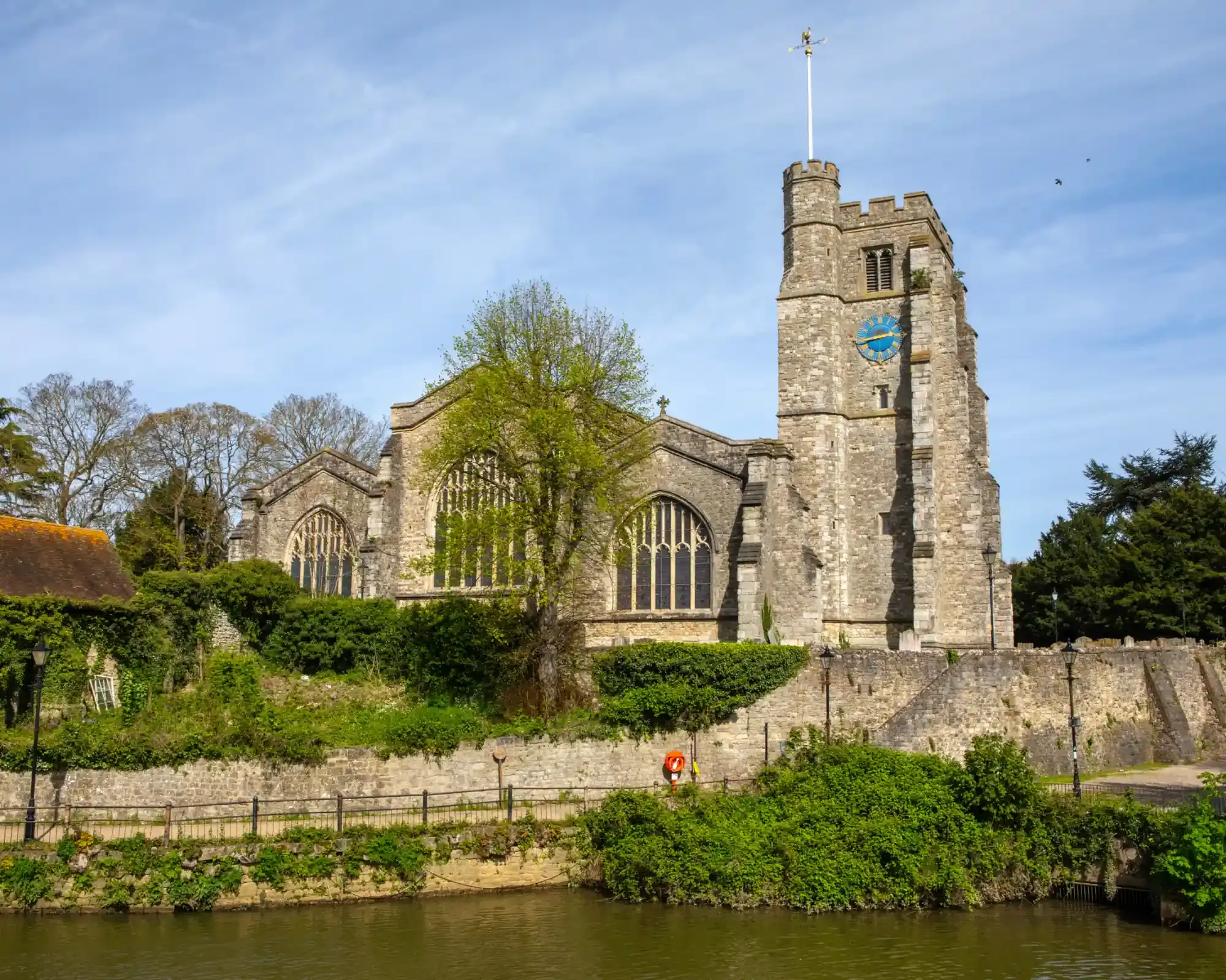 All Saints' Church in Maidstone, a medieval ragstone church with a blue clock face on its tower, beside the River Medway in spring.
