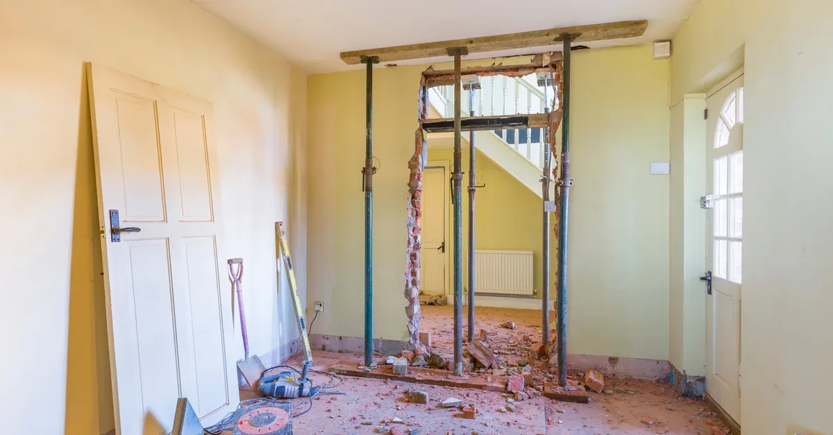 Room in a house being renovated, with a wall partly knocked through and held up by steel props, and bricks and tools on the floor.