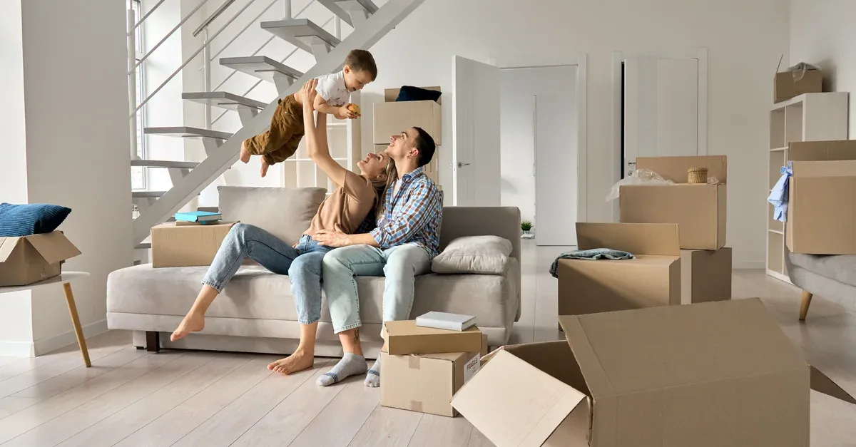 Family relaxing on a sofa in their new home, surrounded by moving boxes, with a parent lifting a laughing toddler into the air.