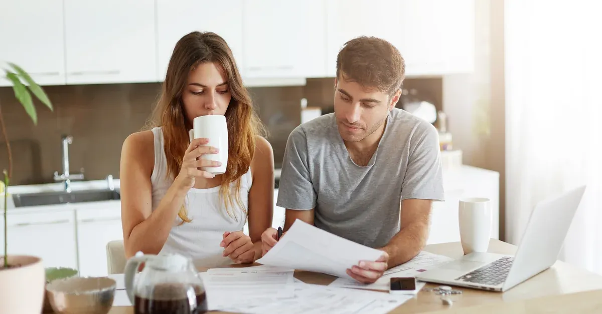 Young couple at a kitchen table looking through financial paperwork with a laptop, working out their house deposit.