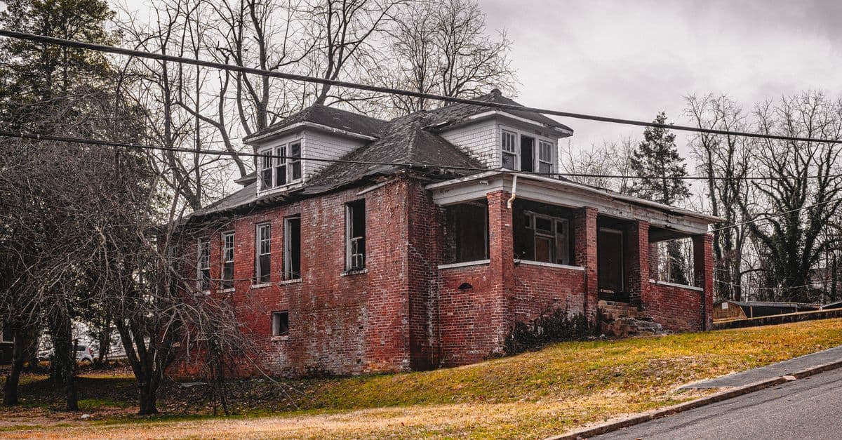 An old abandoned brick house stands amidst bare trees on a cloudy day.