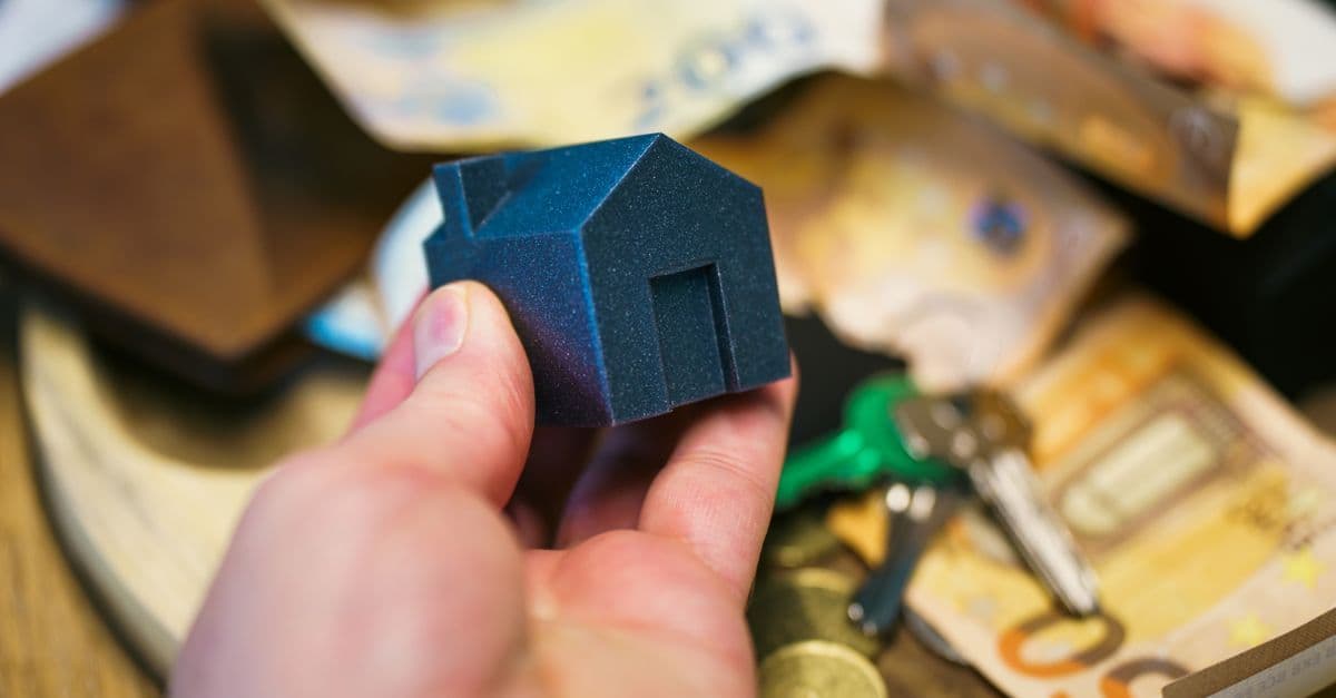 Close-up of a hand holding a small house model surrounded by euro banknotes and keys, symbolizing real estate investment.