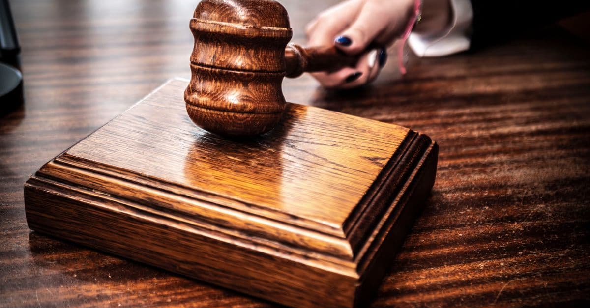 Close-up image of a wooden gavel held by hand on a desk in a courtroom setting.
