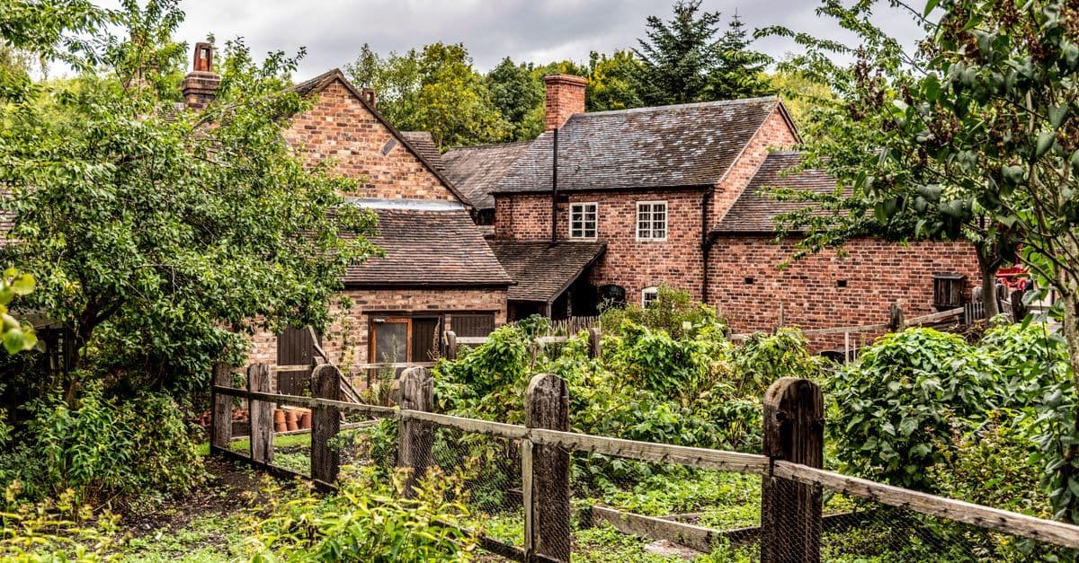 Quaint English brick cottage surrounded by a lush green garden with wooden fencing.