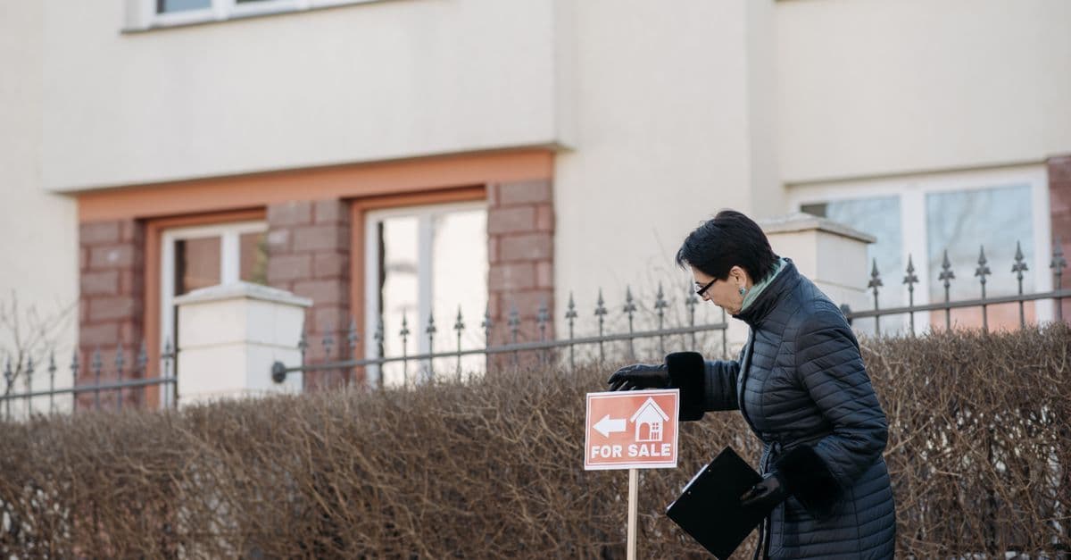 Real estate agent in a black coat placing a 'For Sale' sign in front of a house.