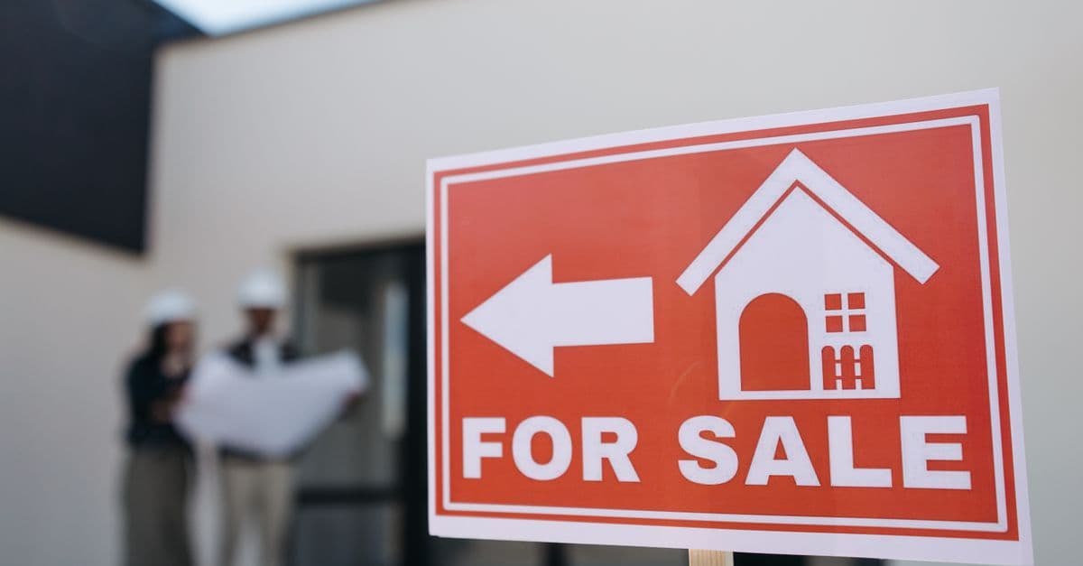 A real estate sign indicates a property for sale as two agents in hard hats discuss building plans outdoors.