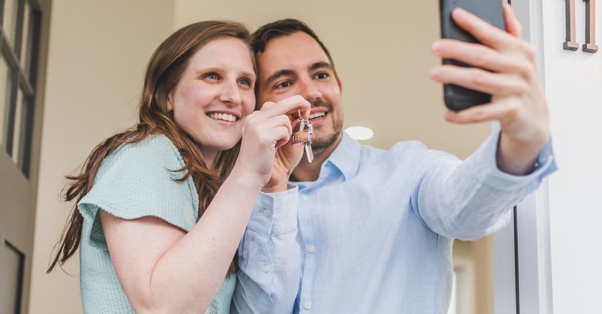 Joyful couple taking a selfie while holding keys to their new home, celebrating a milestone.