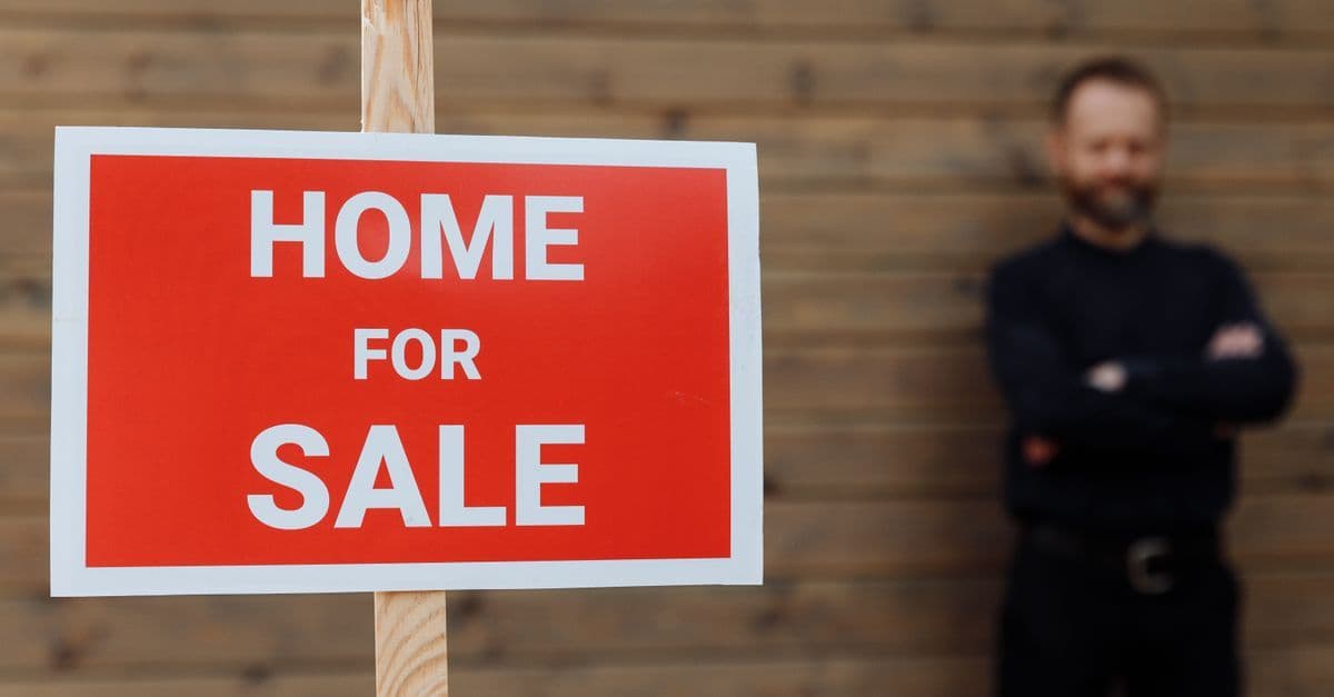 A confident real estate agent stands by a home for sale sign against a wooden wall.
