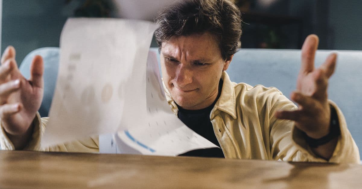 Stressed man throwing documents in frustration at a wooden desk in an indoor setting.