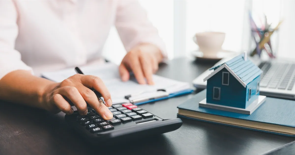 Man sitting at a desk with paperwork, a calculator and a laptop with a house model on a notebook.
