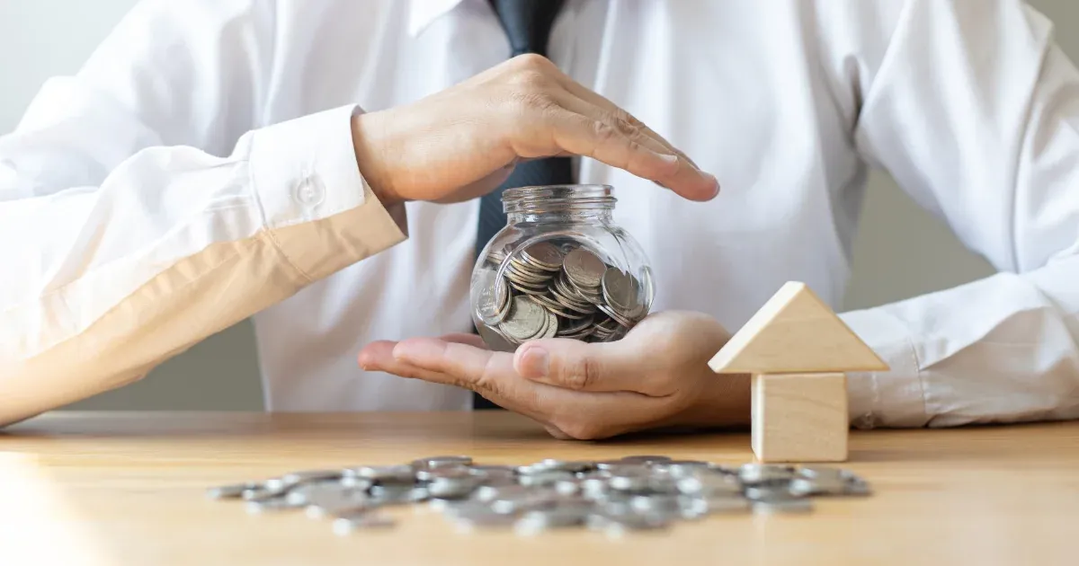 Man in suit sitting at a table with a model house and a jar of money