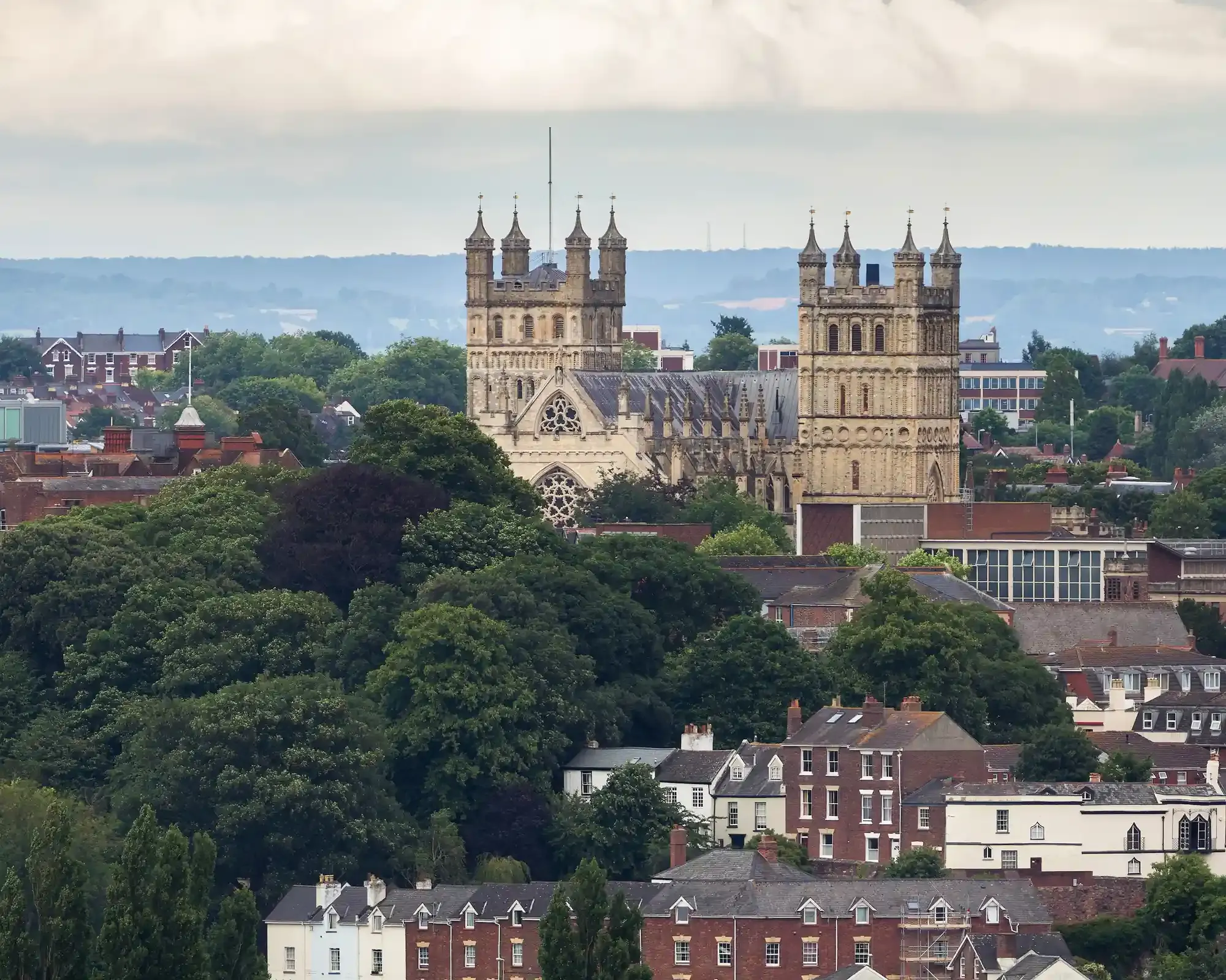 Exeter Cathedral's twin towers rising above trees and red-brick houses, with hills in the distance under a cloudy sky.