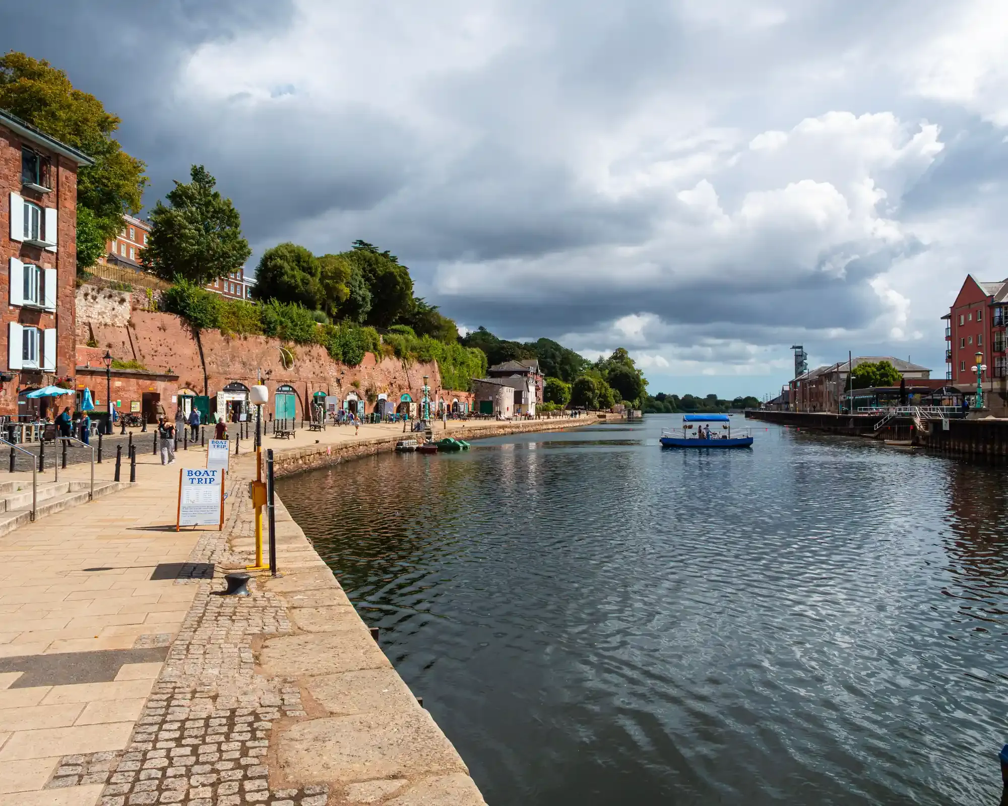 Exeter Quayside under a dramatic cloudy sky, with red sandstone buildings and cellars along the River Exe, and a small ferry crossing the water.