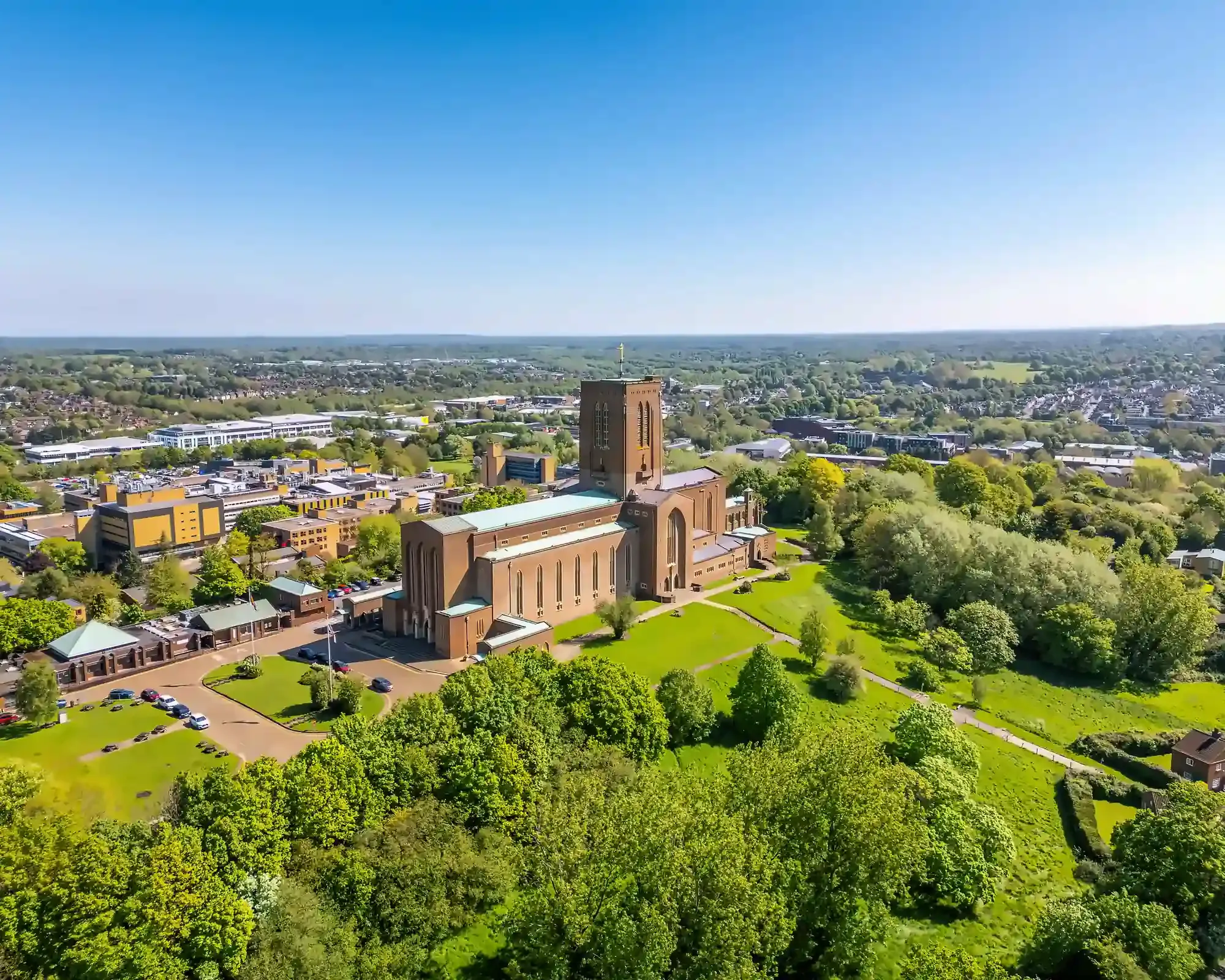 Aerial view of Guildford Cathedral on a sunny day, surrounded by trees and green lawns, with the town and countryside beyond.