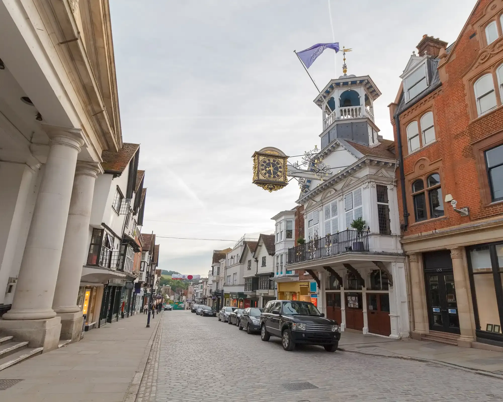 Guildford's cobbled High Street, with the Guildhall's ornate gold clock hanging over the road and historic shopfronts on either side.
