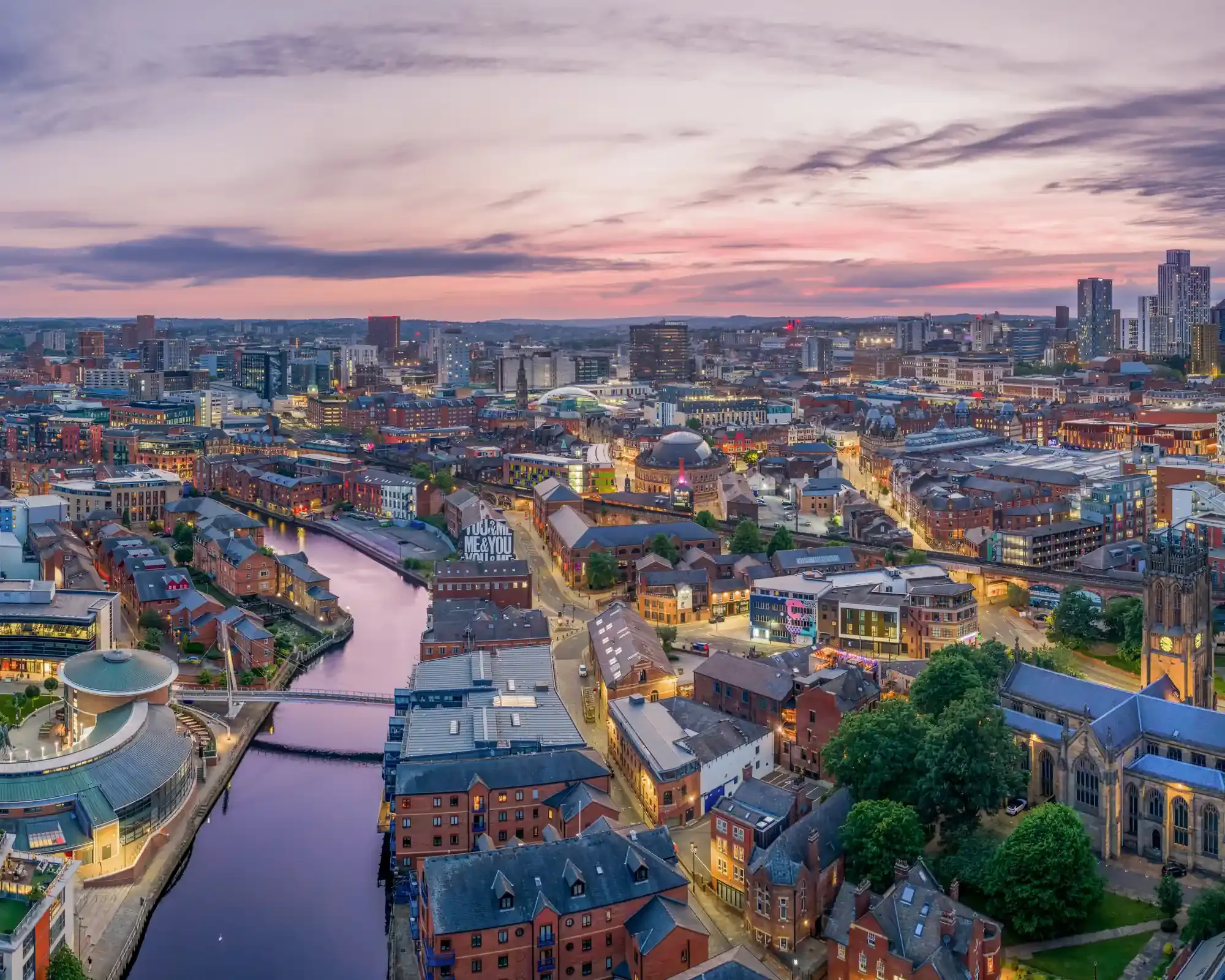 Aerial view of Leeds at dusk, with the River Aire winding through the city centre towards Leeds Minster under a pink and purple sky.