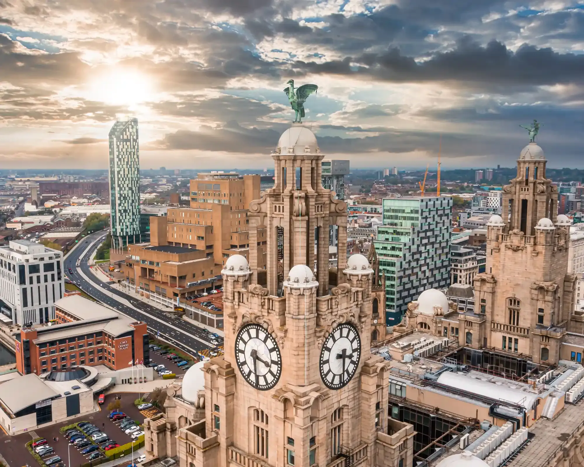 Close aerial view of the Royal Liver Building's clock towers and Liver Birds, with Liverpool's skyline under a dramatic cloudy sky.